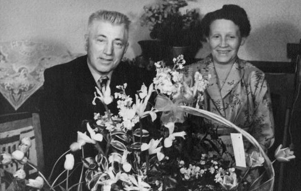 Twee mensen zitten achter een groot bloemstuk, poseren binnen en lachen naar de camera.