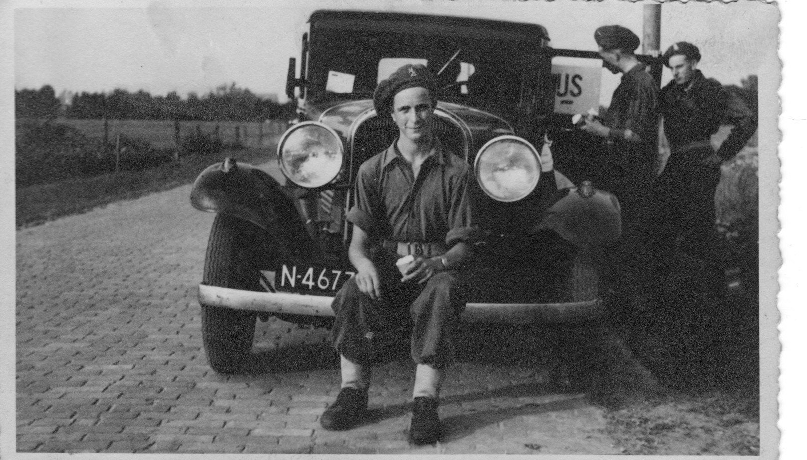 Zwart-witfoto van een oldtimer met twee mannen die op de weg poseren, waarvan er één op de bumper zit.