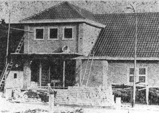 Zwart-witfoto van een klein huis in aanbouw met steigers en een gedeeltelijk afgewerkte veranda.