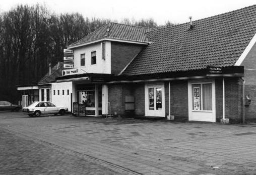 Zwart-witfoto van een klein gebouwtje langs de weg met een geparkeerde auto en een beboste achtergrond.
