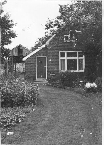 Klein bakstenen huisje met een voordeur, raam en een gebogen zandpad, omgeven door bomen en struiken.