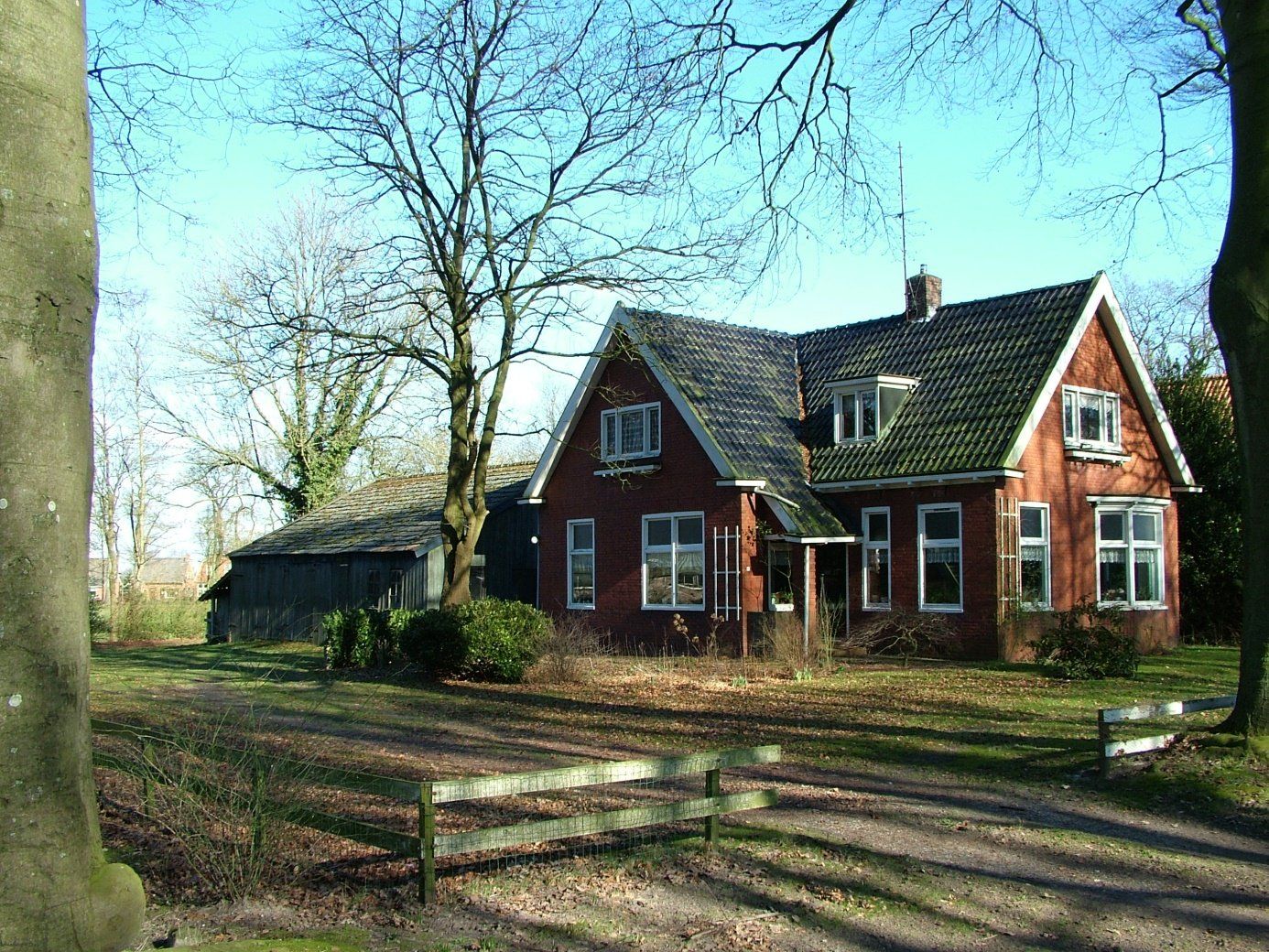Een rood bakstenen huis met een steil dak, gelegen naast kale bomen en een omheinde tuin in een zonnige, landelijke omgeving.
