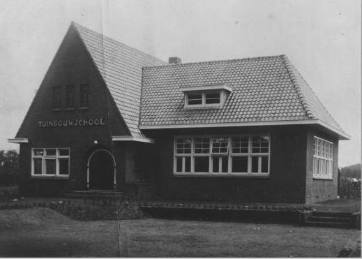Zwart-witfoto van een klein bakstenen schoolgebouw met een steil dak en een bord boven de ingang.