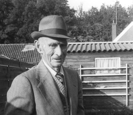 Een man met een fedora en een pak staat buiten voor een gebouw en bomen en kijkt in de camera.