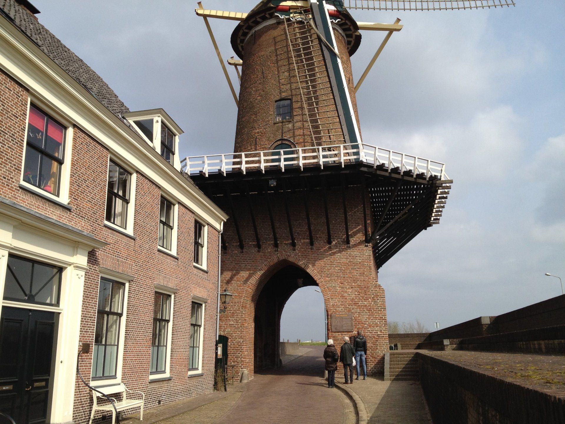 Een bakstenen windmolen met witte leuningen staat naast een kanaal, van onderaf gezien, met een paar mensen die er vlakbij lopen.