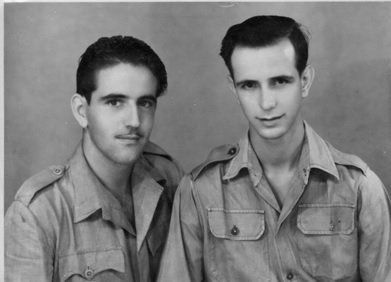 Twee mannen in lichte, militaire shirts poseren tegen een effen achtergrond.