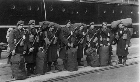 Soldaten met geweren en plunjezakken gaan aan boord van een schip in een kade, in zwart-witfoto.