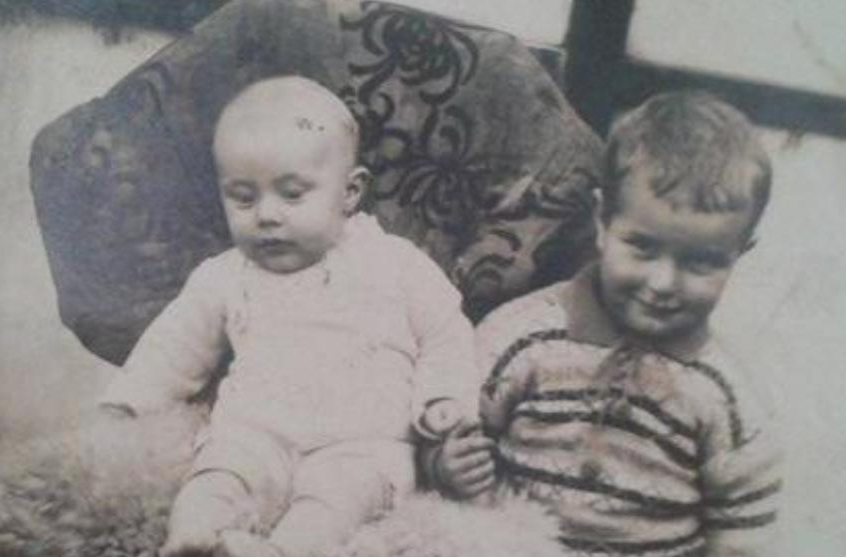 Twee kleine kinderen zitten op een stoel met een patroon, de ene kijkt naar beneden en de andere lacht naar de camera.