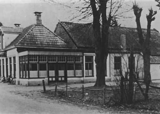 Zwart-witfoto van een klein paviljoenachtig gebouw met bomen en een uithangbord ervoor.