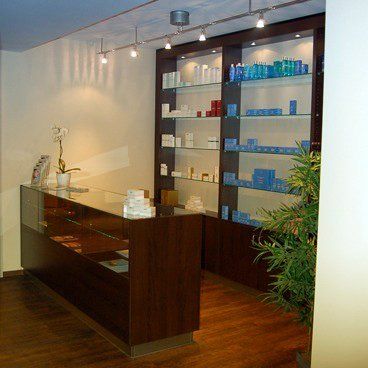Empfangsbereich in einem Spa. Theke und Regale aus dunklem Holz mit Produktpräsentation, Deckenbeleuchtung und einer Pflanze.