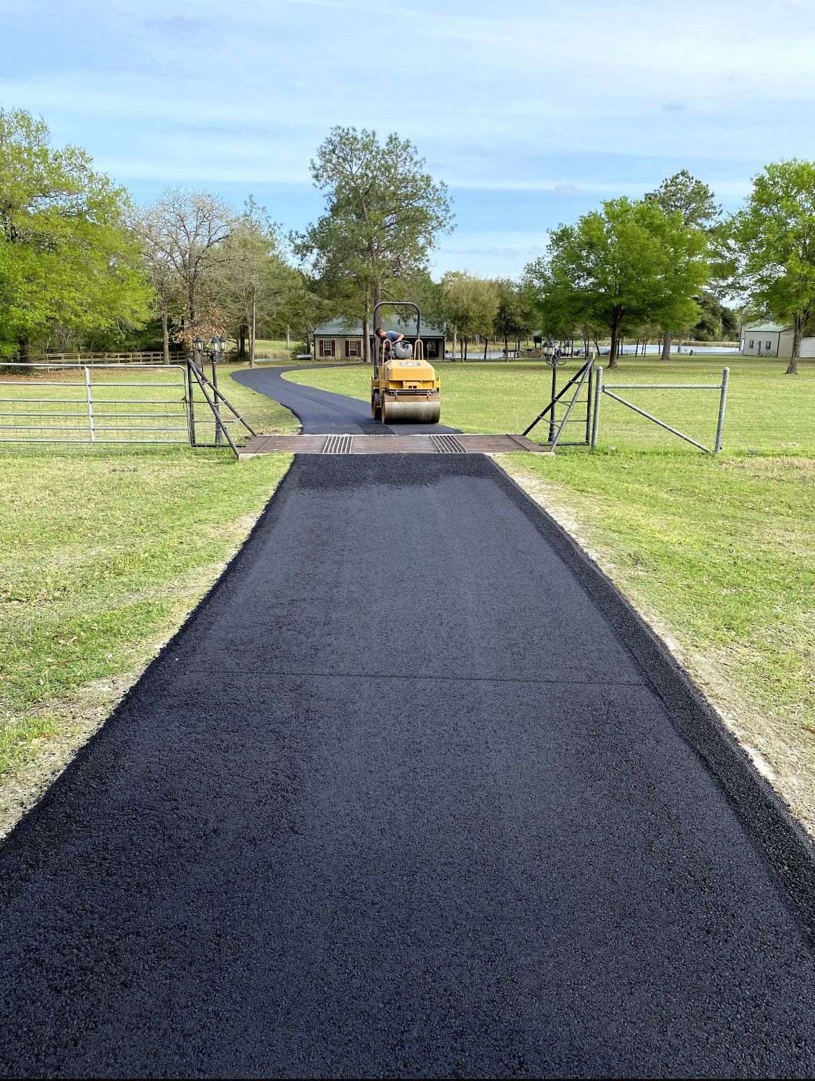 Fresh black asphalt path leading to a yellow roller and open gate in a grassy park