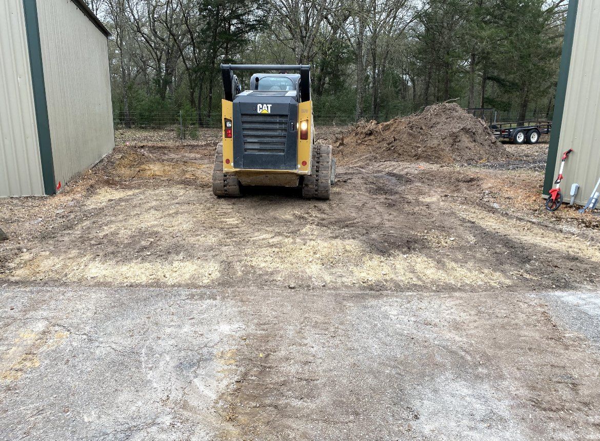 Yellow skid steer in a gravel lot between two buildings, with dirt piles in the background.