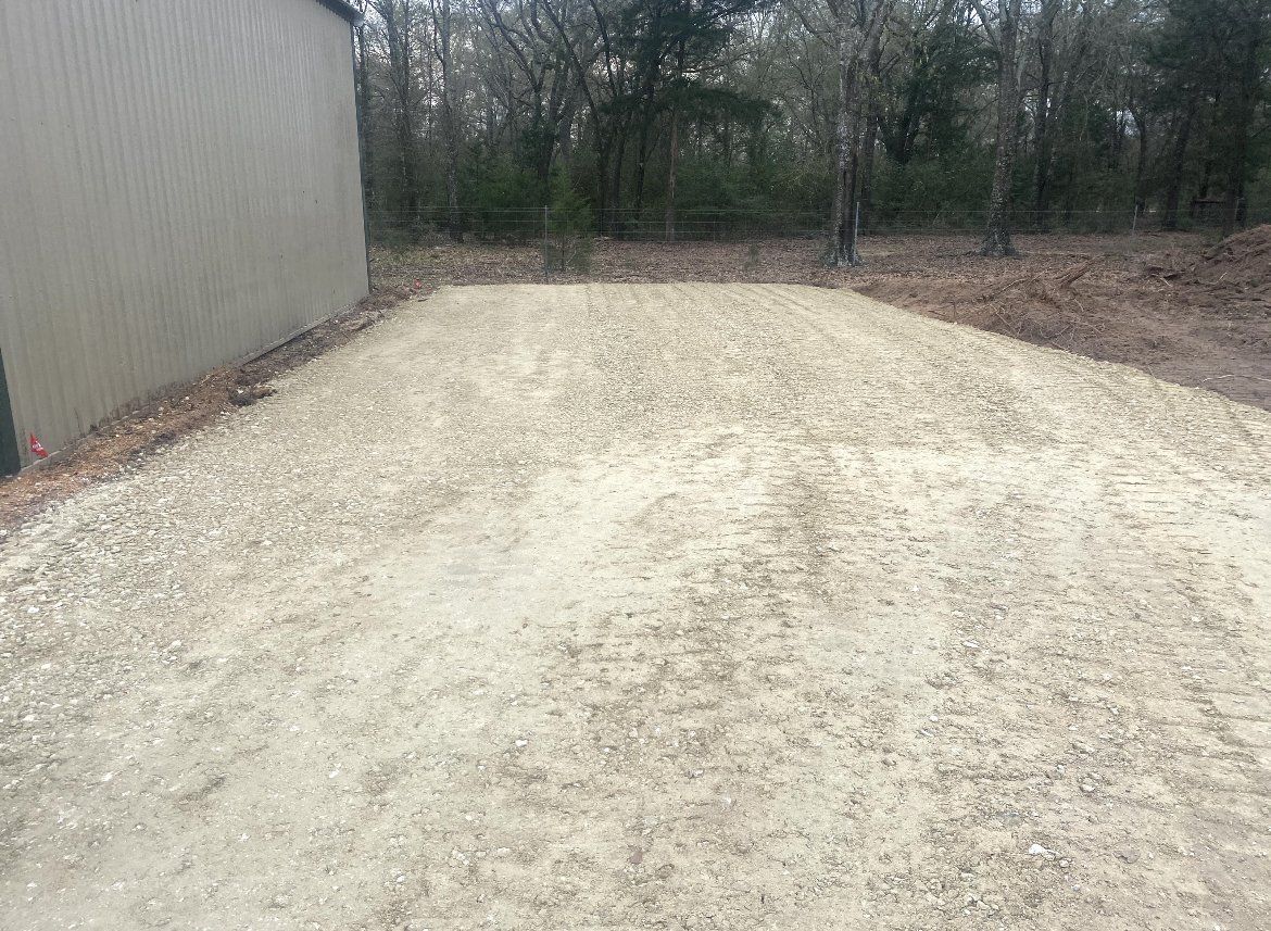 Gravel driveway beside a corrugated metal building, leading into a wooded area