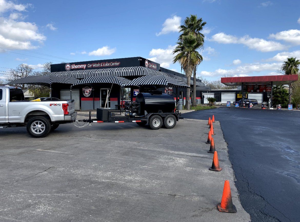 Outdoor parking lot with orange cones, a silver pickup, and a black trailer near a storefront under a blue sky