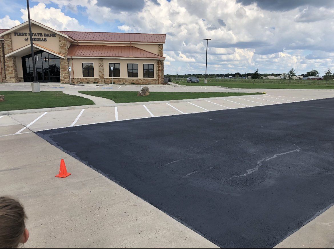 Empty parking lot beside a tan building under a cloudy sky, with orange cones marking the pavement.