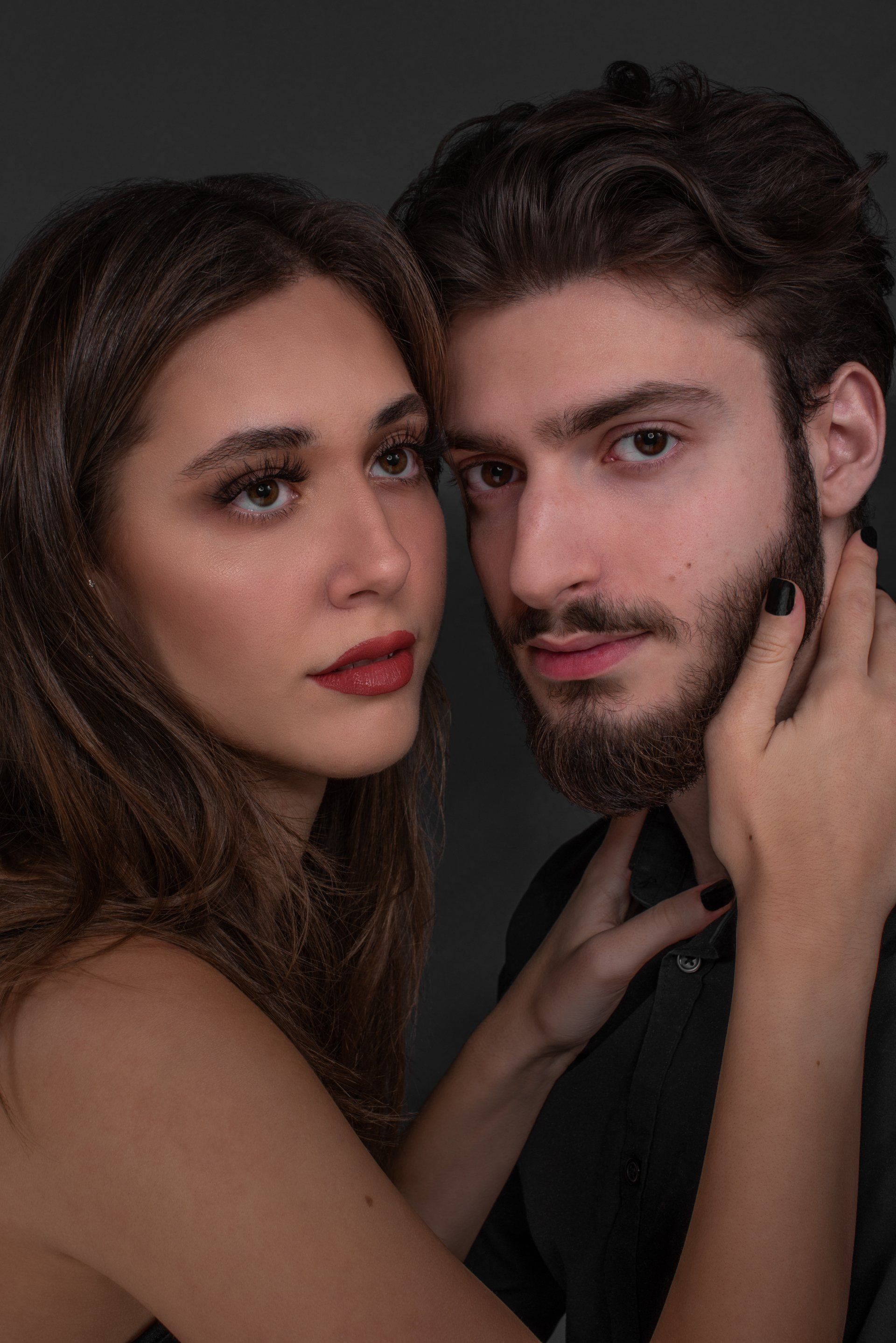 Woman with dark hair touches a man's face; both look at the camera with serious expressions, against a dark background.