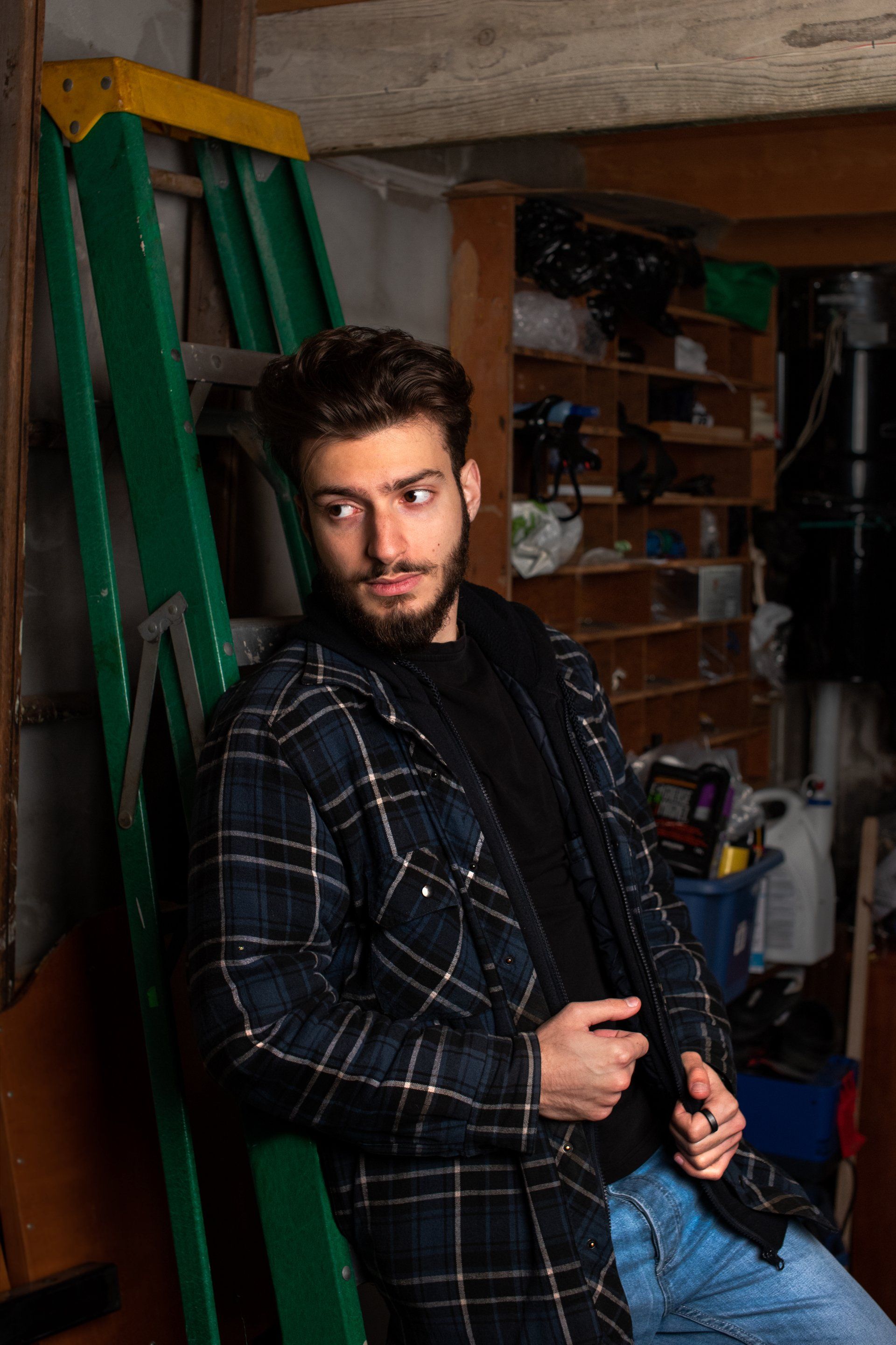 Man leaning against a green ladder in a cluttered shed, wearing a plaid shirt and looking off to the side.