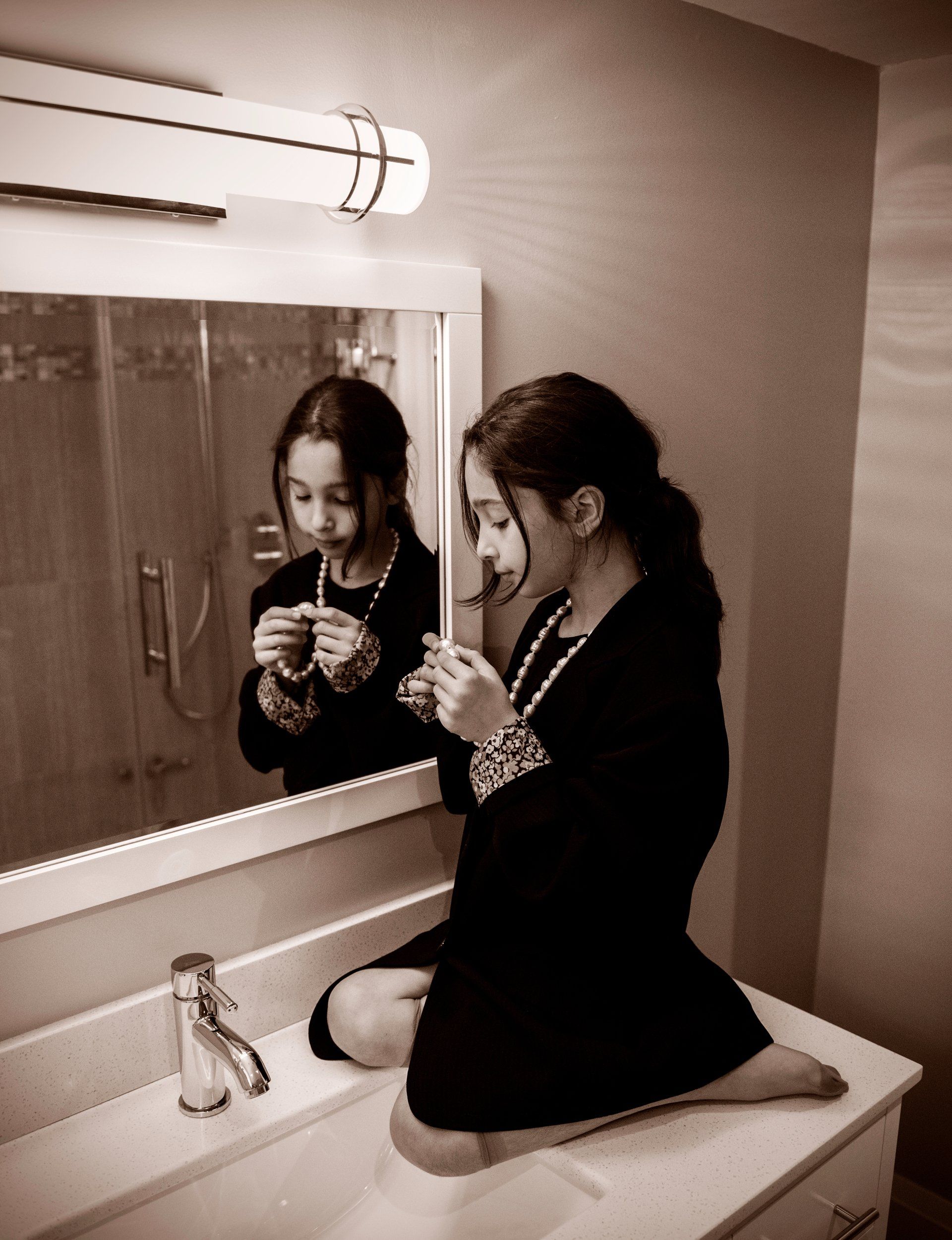 Girl in dark outfit, sitting on bathroom sink, looking at jewelry. Reflection in mirror.
