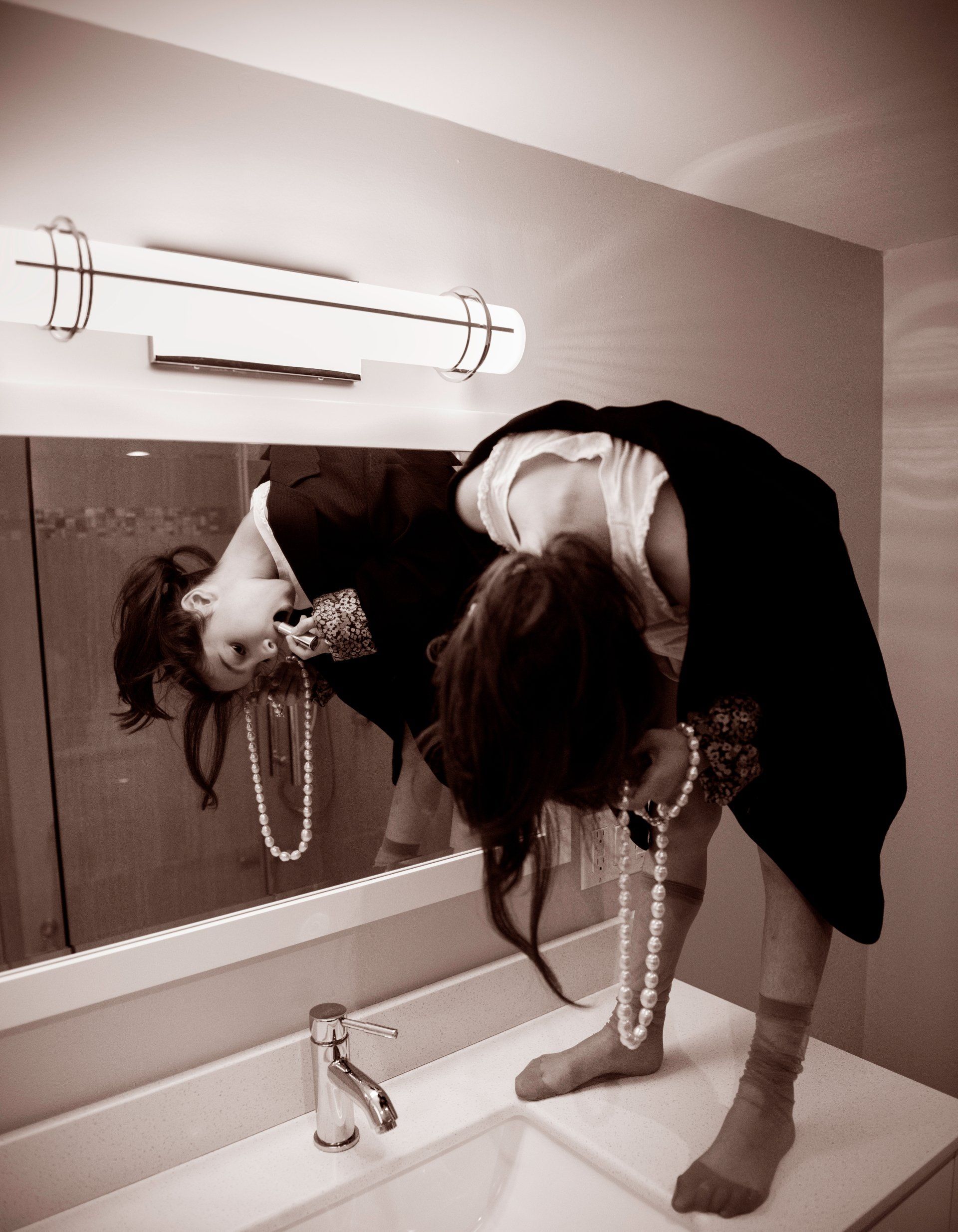 Woman bending over a bathroom sink looking into a mirror, holding pearls, wearing a jacket.