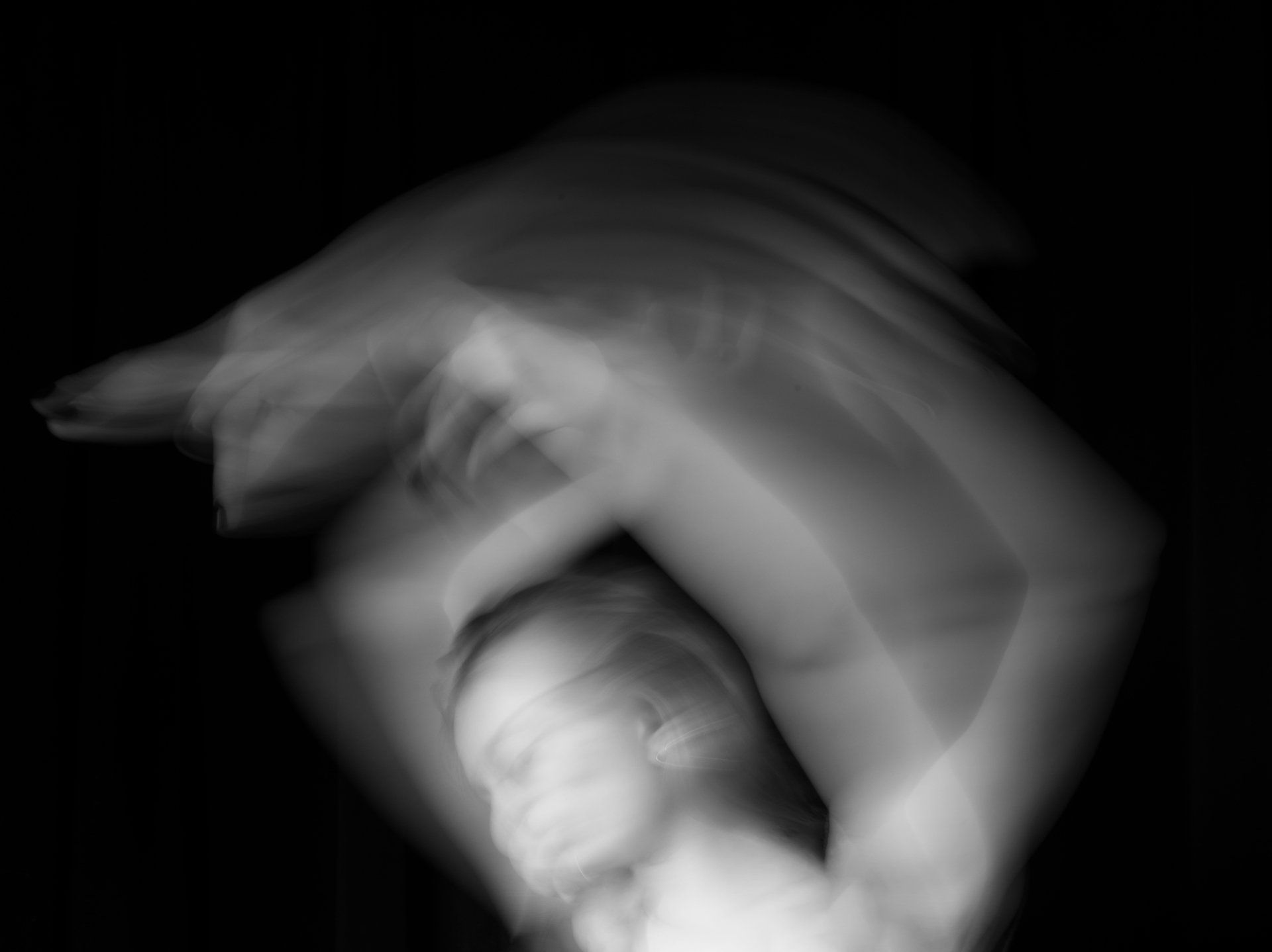 Blurred figure in motion, arms raised above head. Black and white, against a dark background.