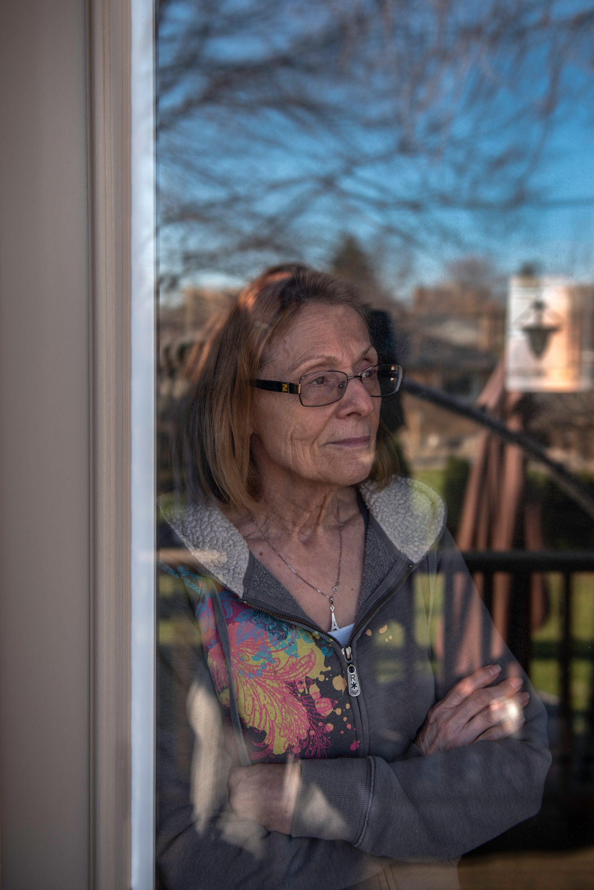 Woman looking out a window with arms crossed, reflecting trees and sky.