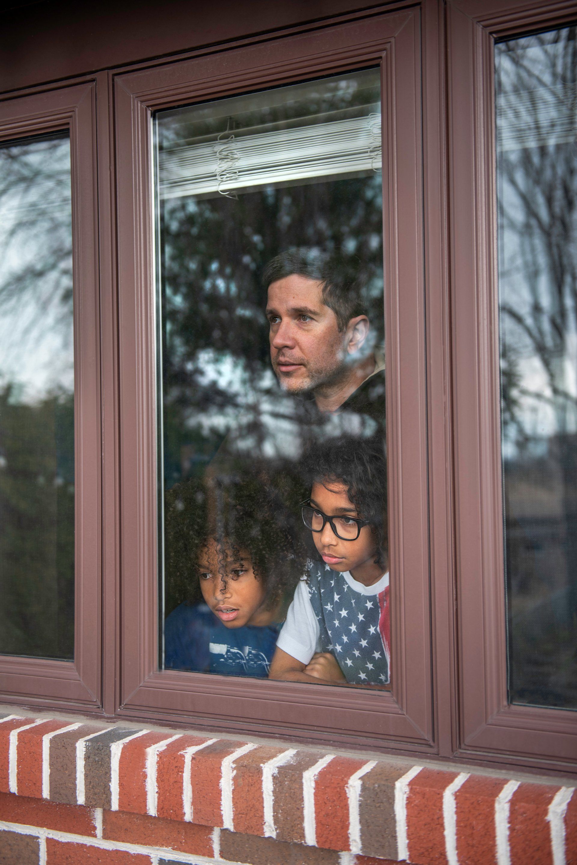 A man and two children look out a window, brown frame and brick exterior visible.
