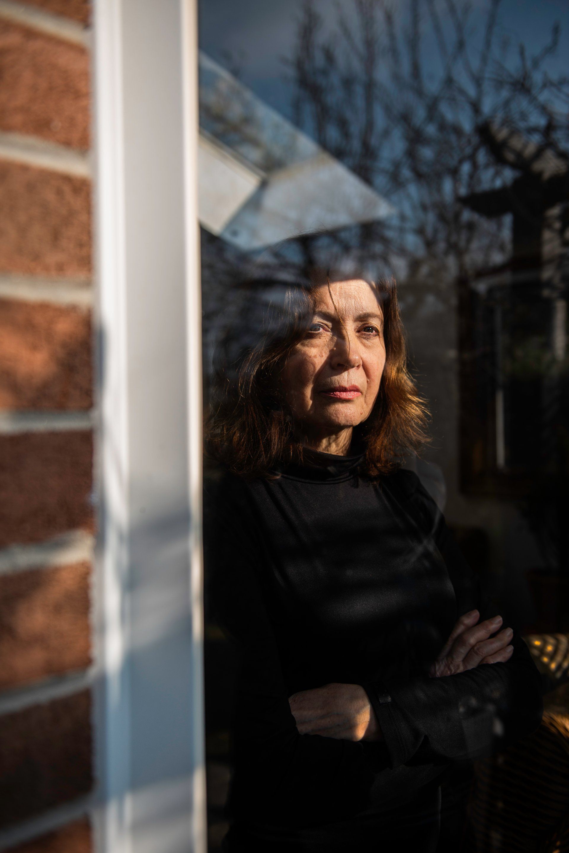 Woman looking out window, arms crossed. Reflected trees visible.