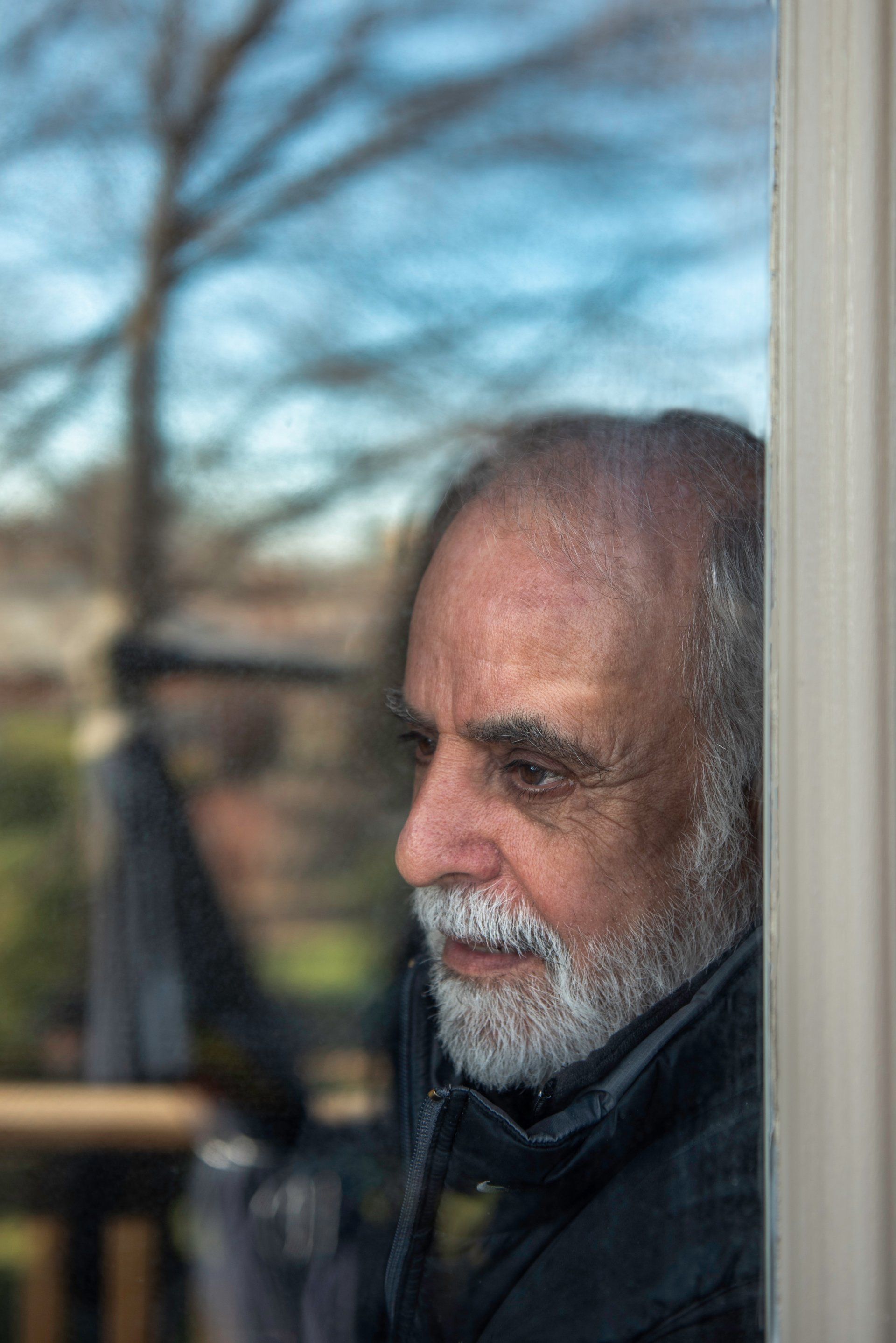 Man with gray beard looking out a window at a blurred outdoor scene with trees and blue sky.