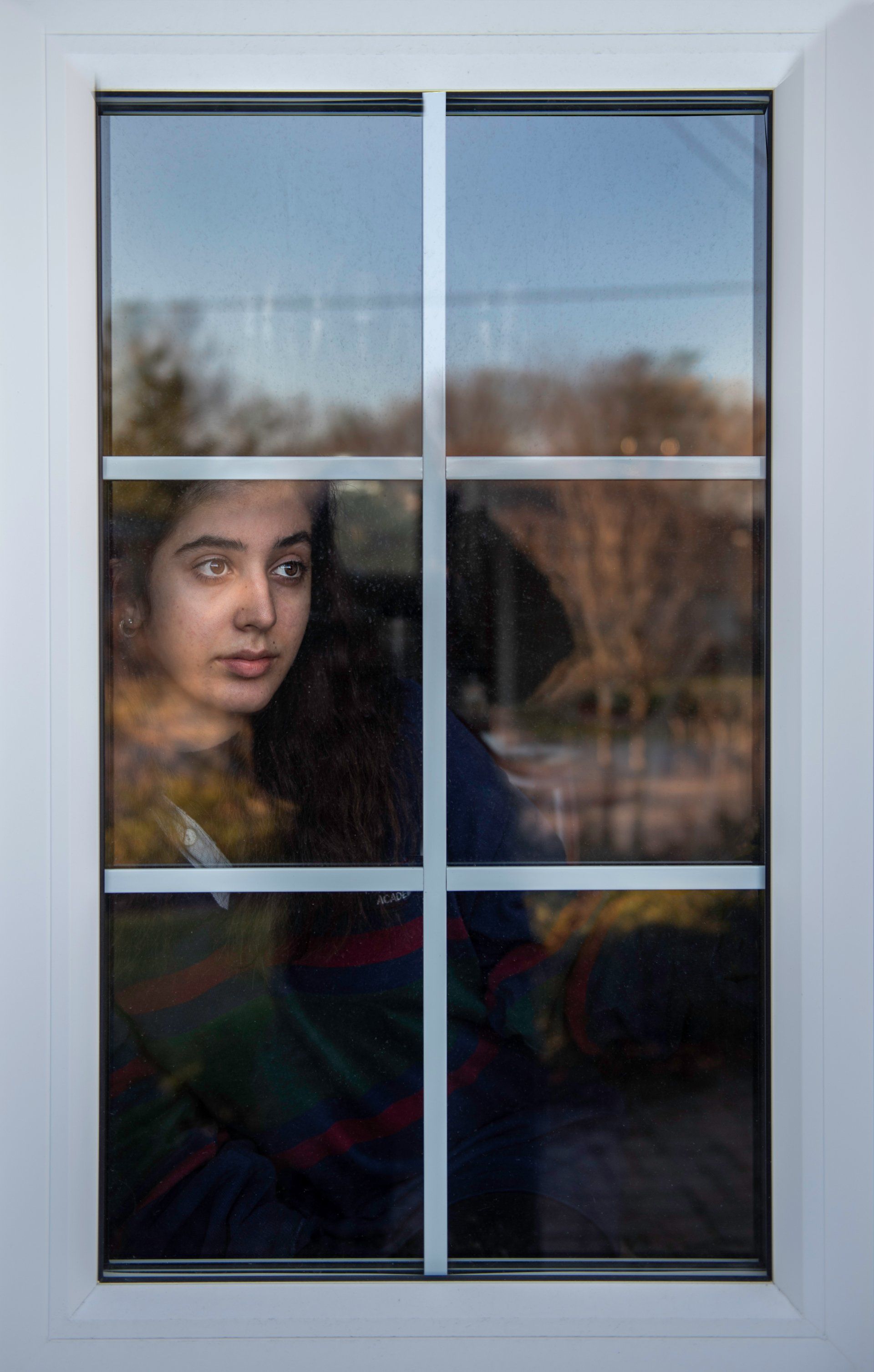 Woman looking out a window. She wears a striped shirt; trees and sky are reflected in the glass.
