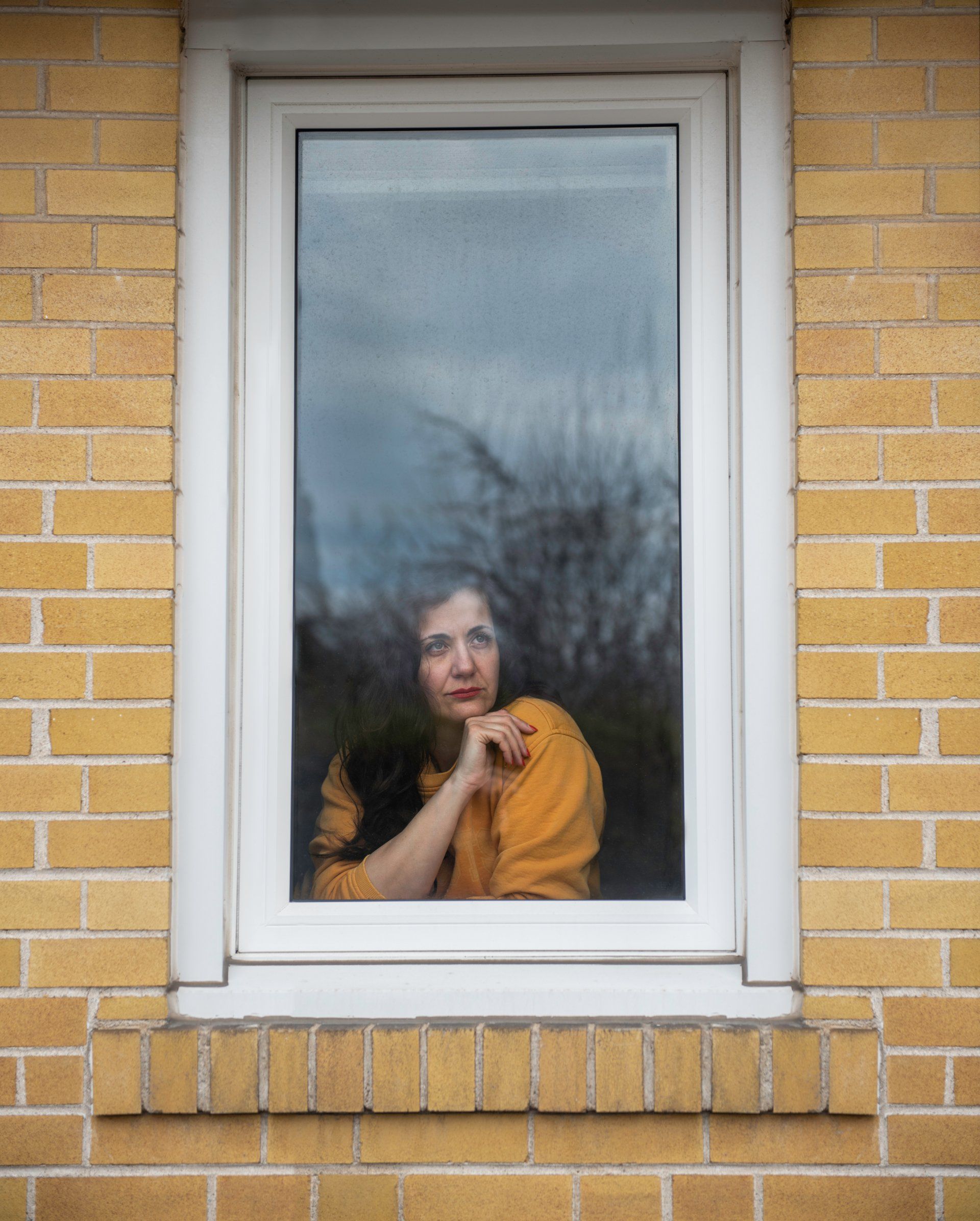 Woman looking out a window. Yellow shirt, brick wall, reflection of trees visible.