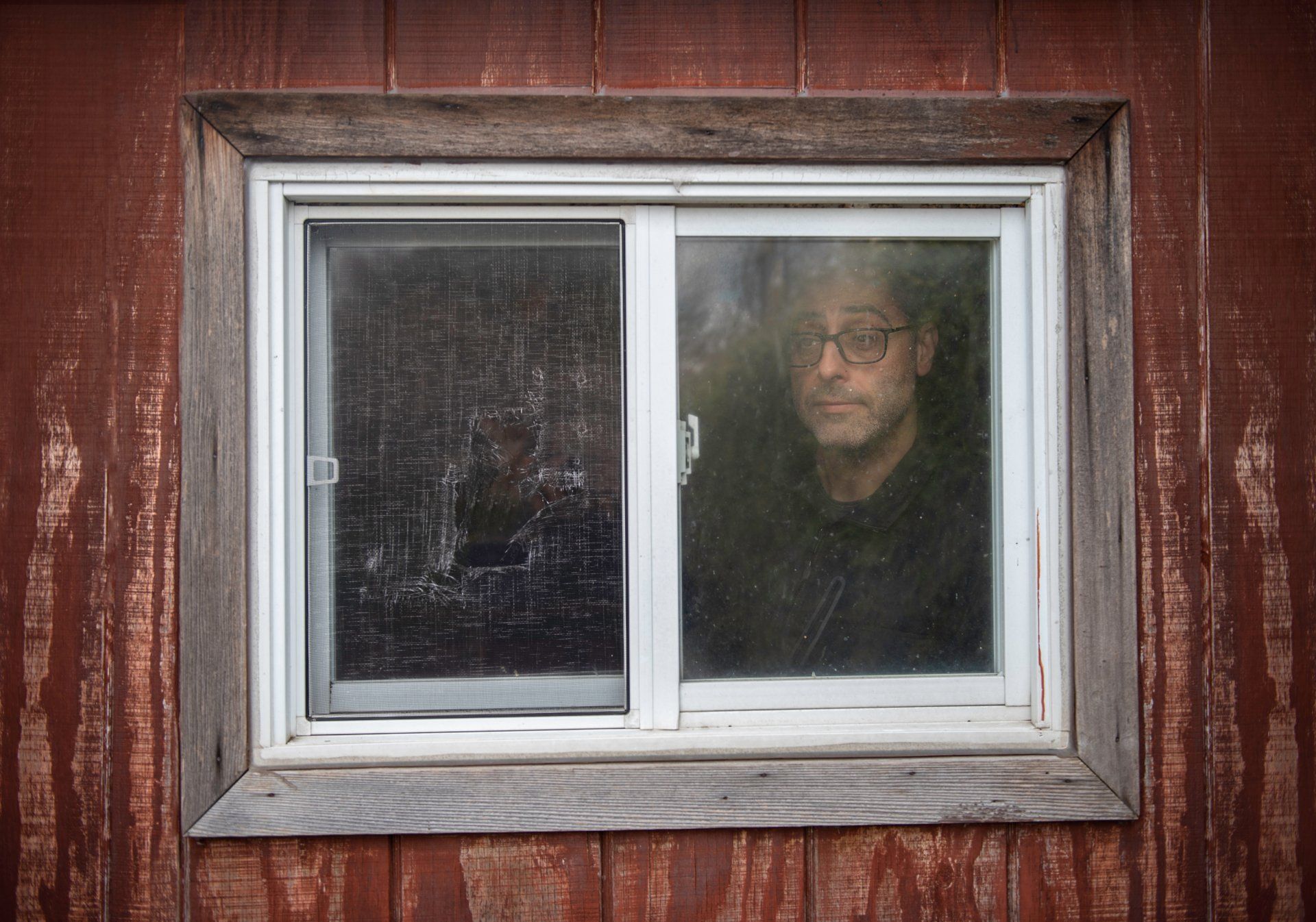 Man looking out a window. Window is set in a red, weathered wooden wall.