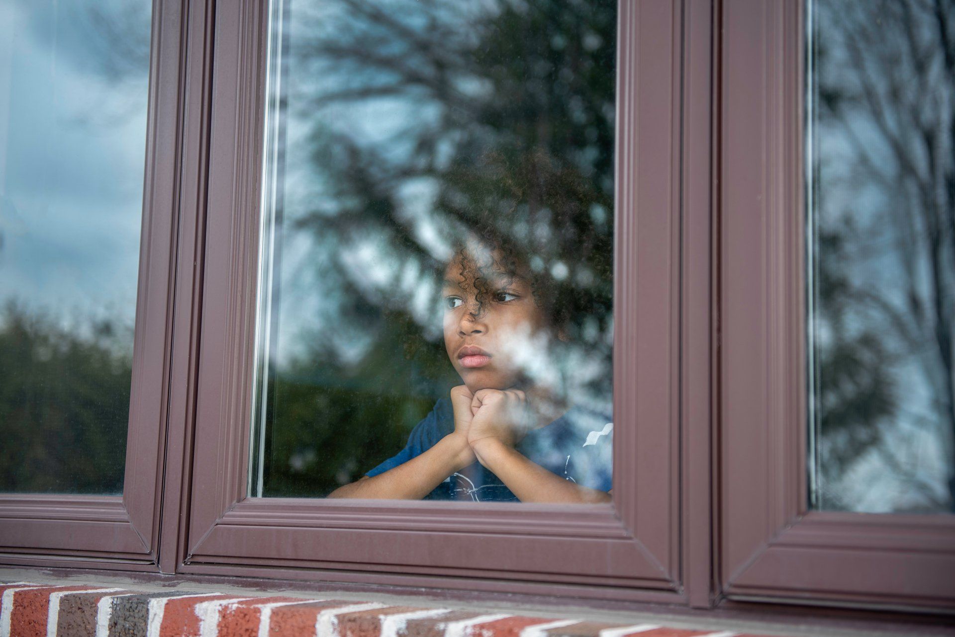A person looking out a window with hands on their chin, brown window frame, brick wall, and trees.