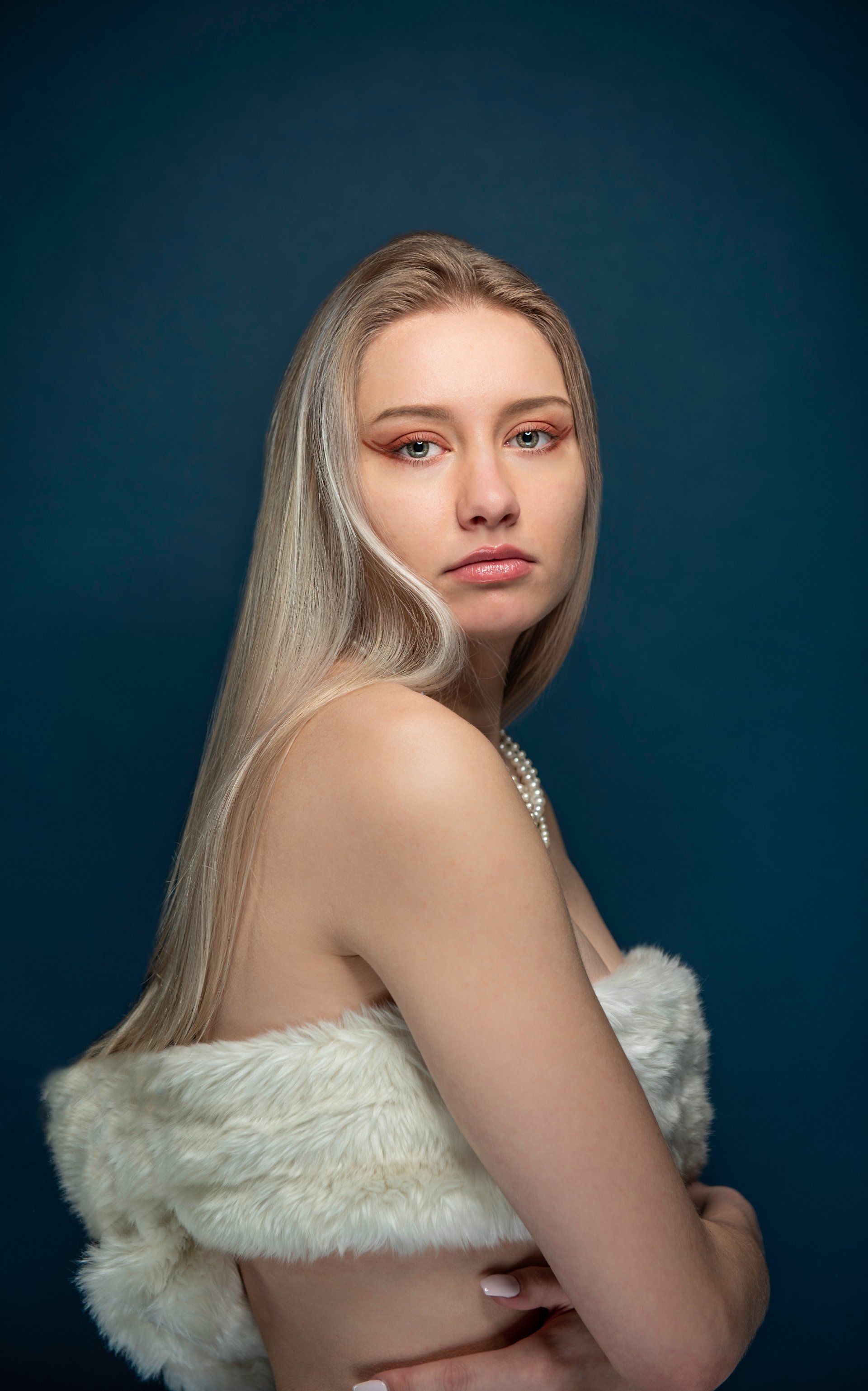 Blonde woman with fair skin, wearing white fur wrap, looking at camera against a blue backdrop.