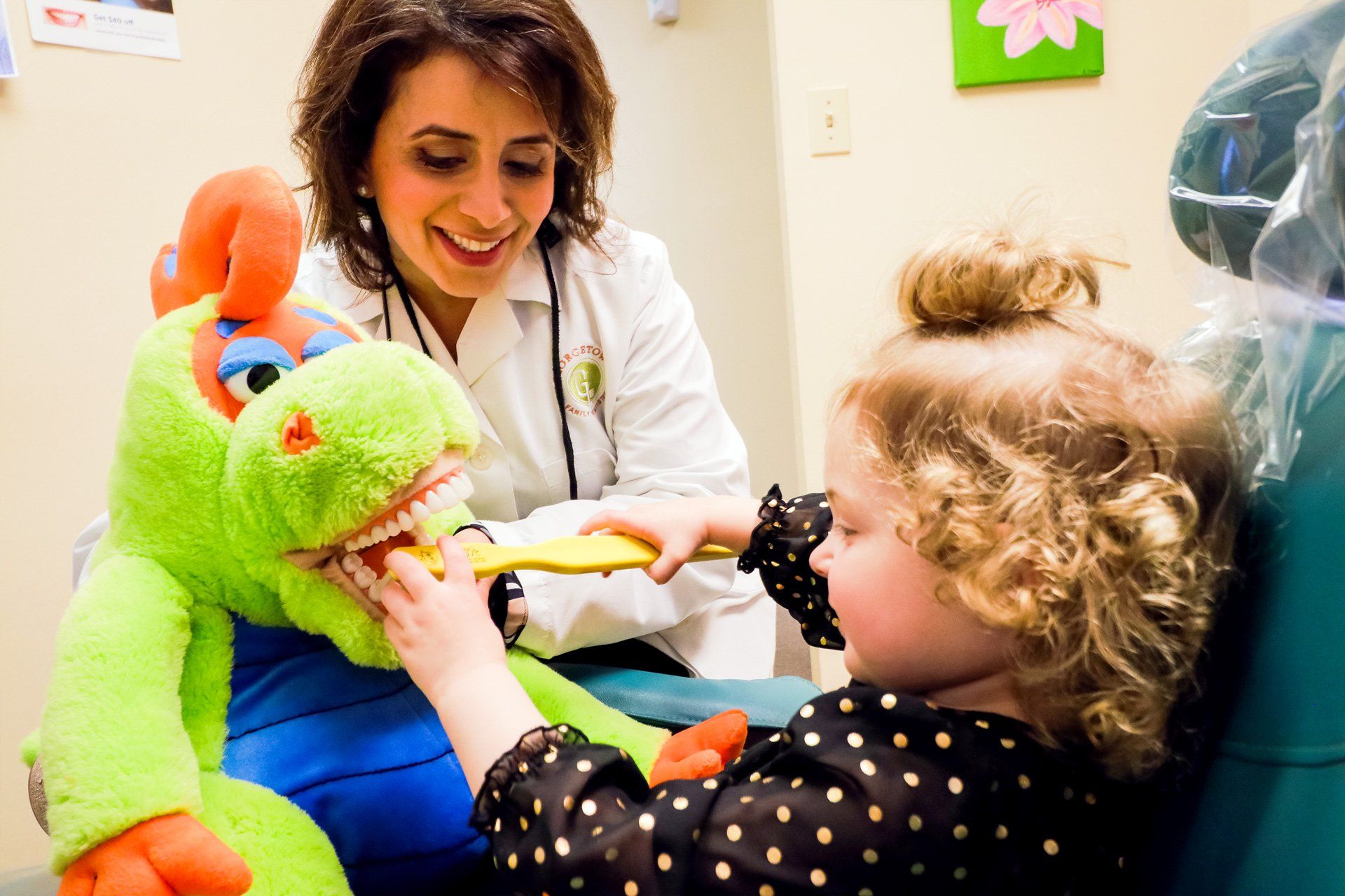 Dentist shows girl how to brush toy dragon’s teeth in a dental office.