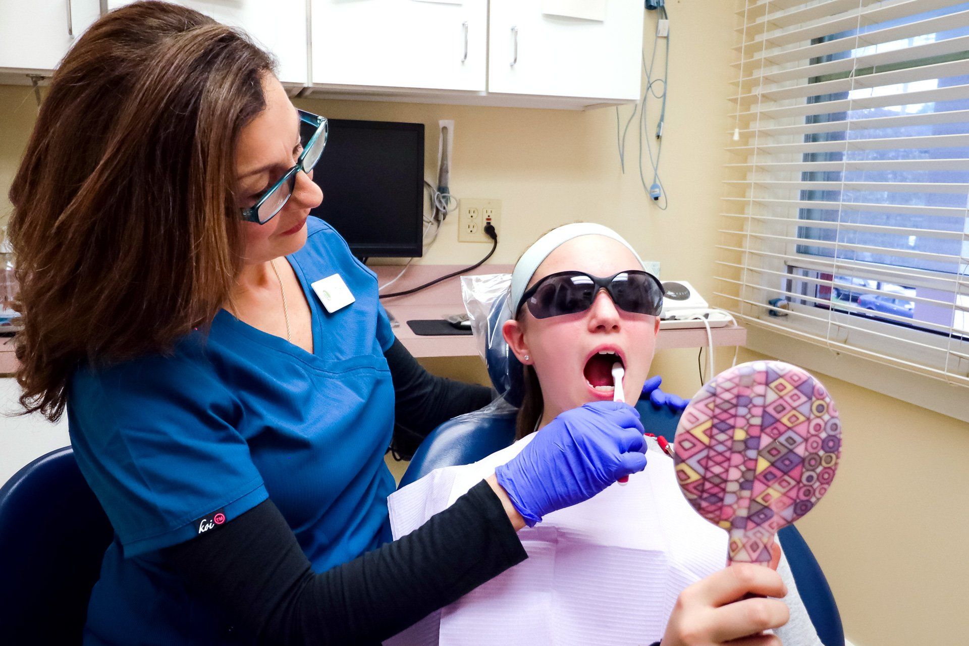 Dentist demonstrating teeth brushing on a young patient in a dental office; both wearing protective eyewear.