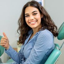 Woman in a blue shirt smiles with a thumbs up while sitting in a dental chair.