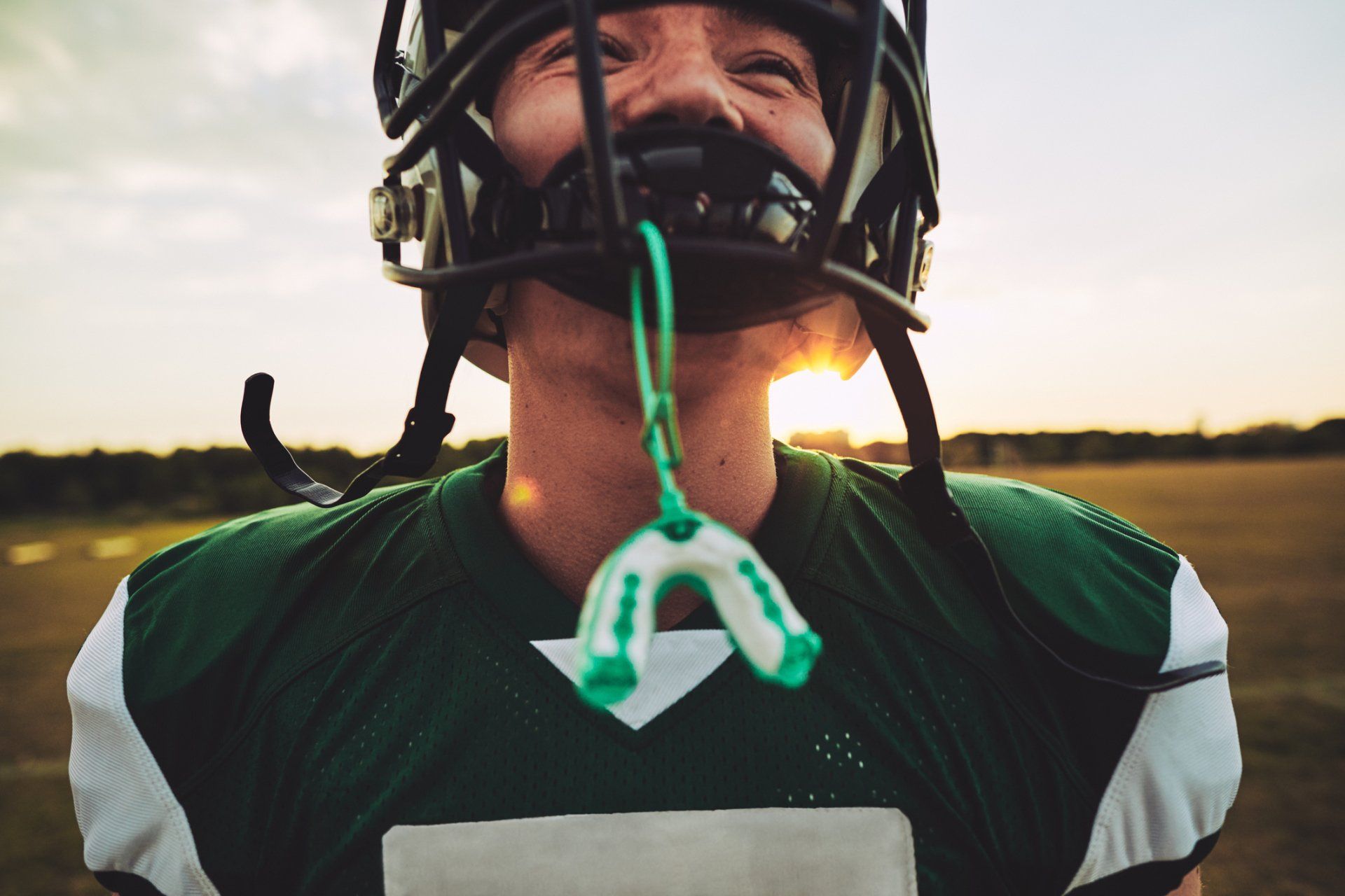 Football player with helmet, green jersey, mouthguard hanging, sun in background.