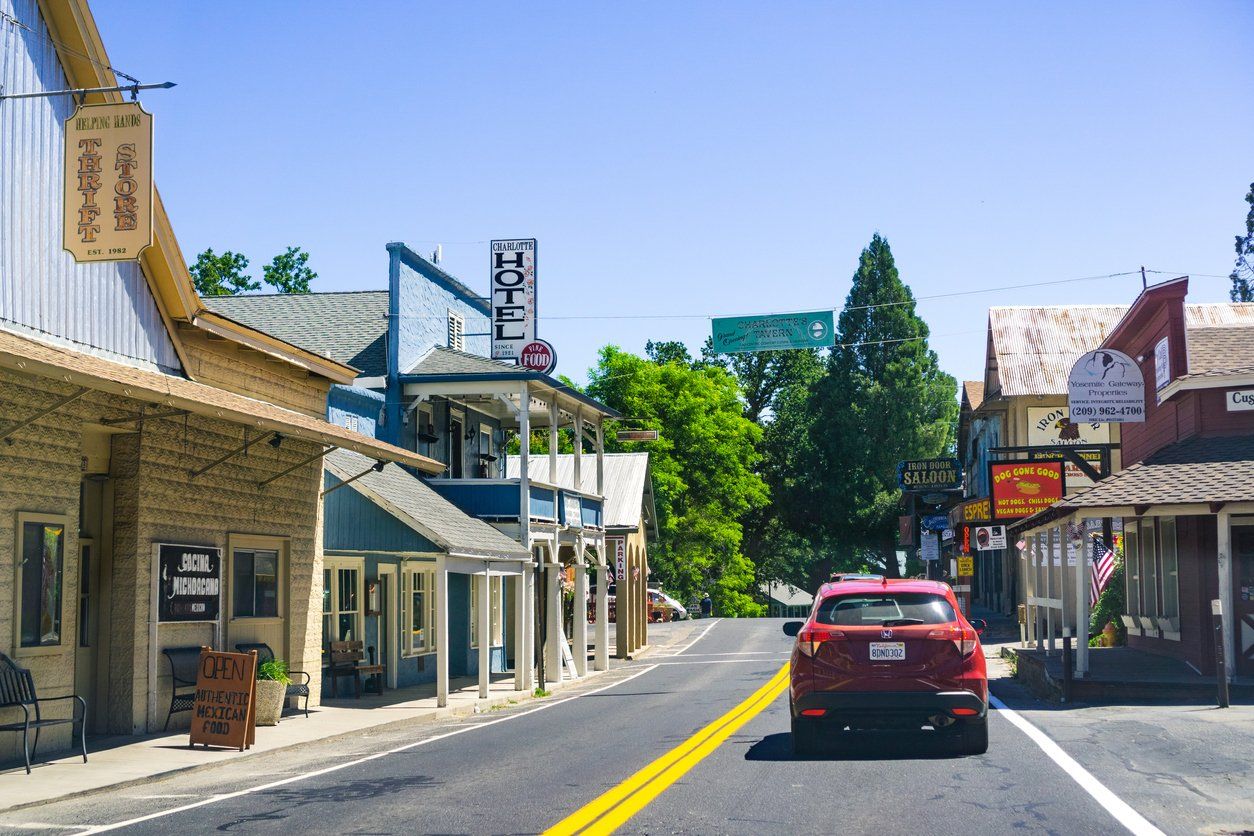 A red car driving down a sunny street lined with colorful buildings in a small town.