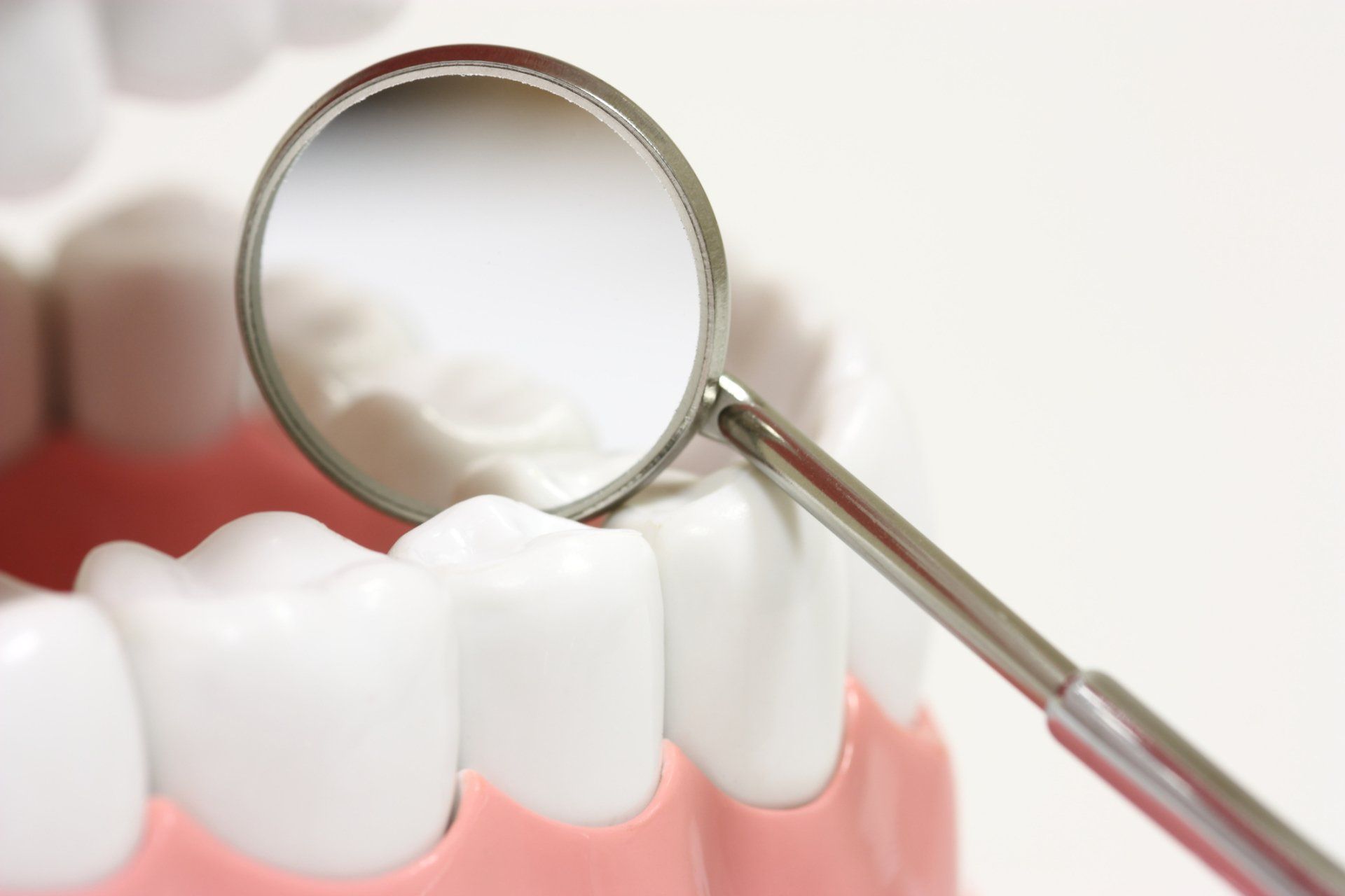 Dentist's mirror inspecting model of white teeth, pink gums, and a white background.