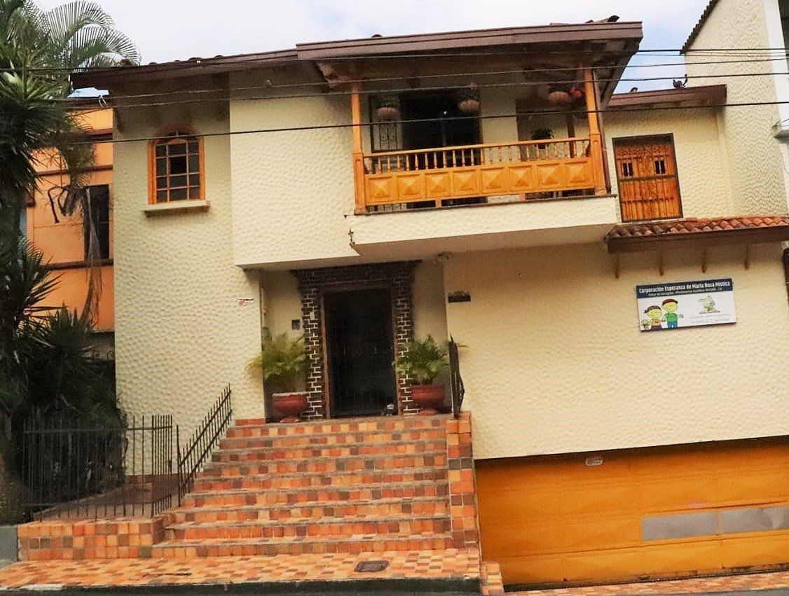 Edificio beige de dos pisos con balcón y entrada de madera, puerta naranja, escalones de ladrillo y un cartel.