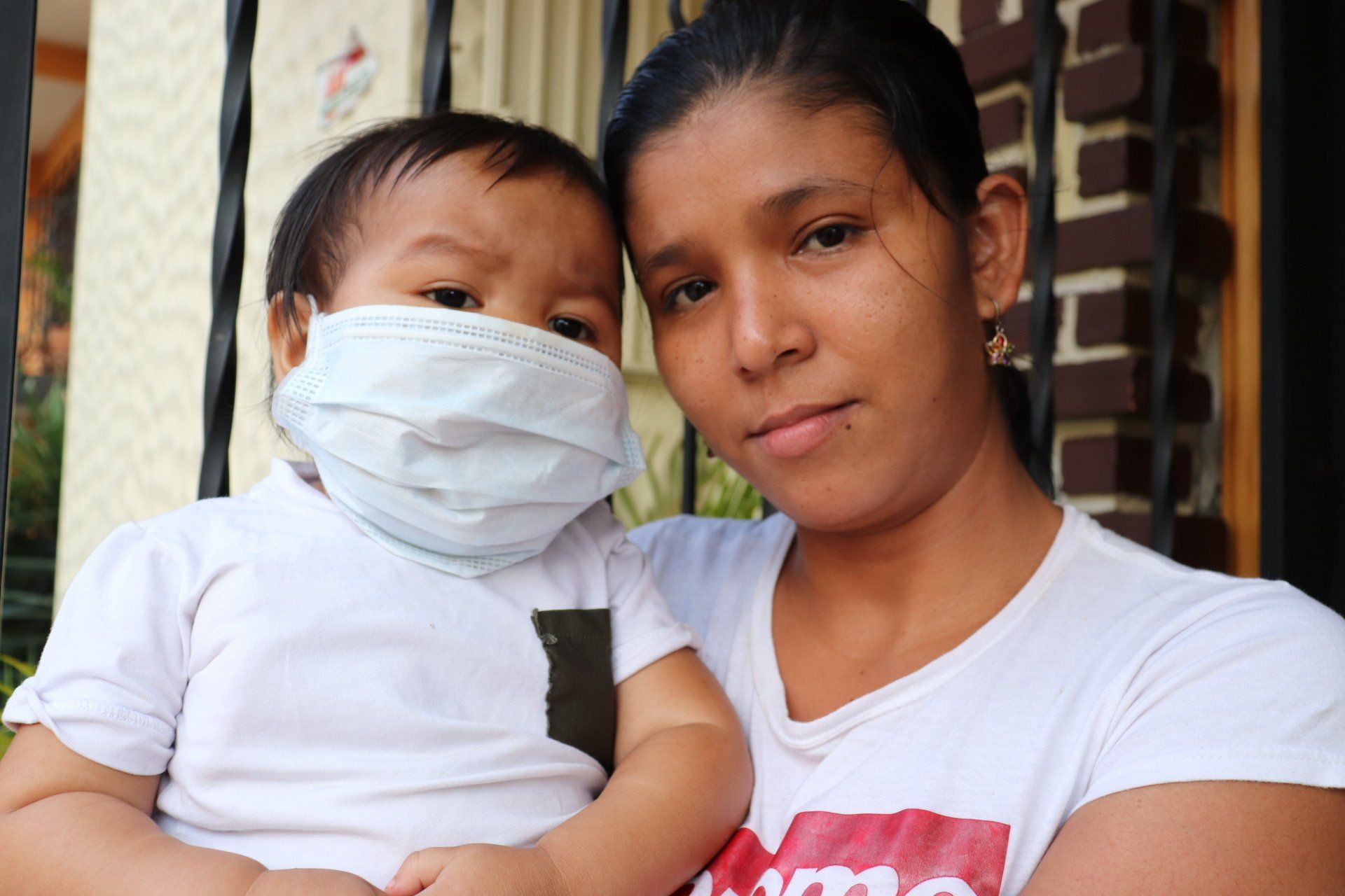 Mujer y niño pequeño, ambos de piel clara, el niño con mascarilla, al aire libre frente a un edificio.