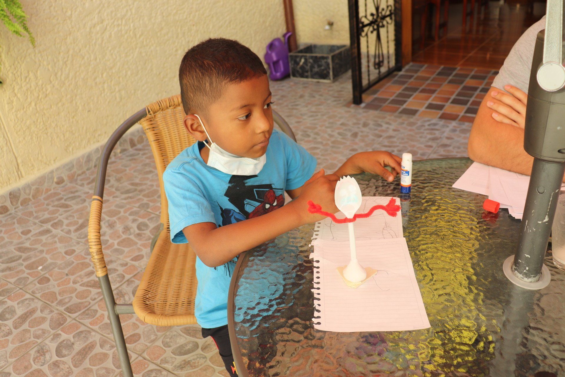 Niño con camisa azul haciendo manualidades en una mesa, trabajando en un proyecto con una cuchara y una barra de pegamento.