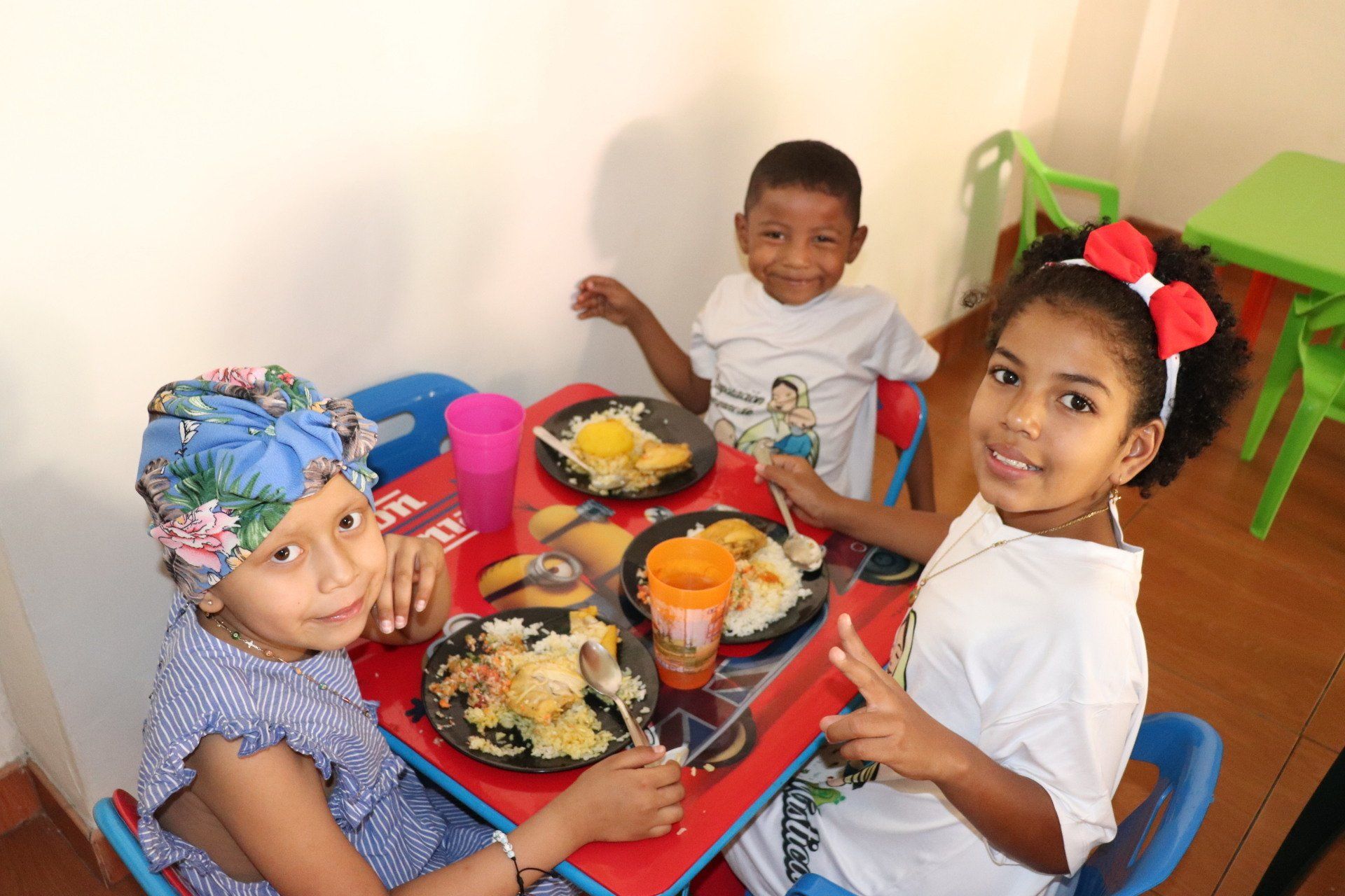 Tres niños comen en una mesa roja. Sonríen, uno con la cabeza cubierta y el otro haciendo el signo de la paz.