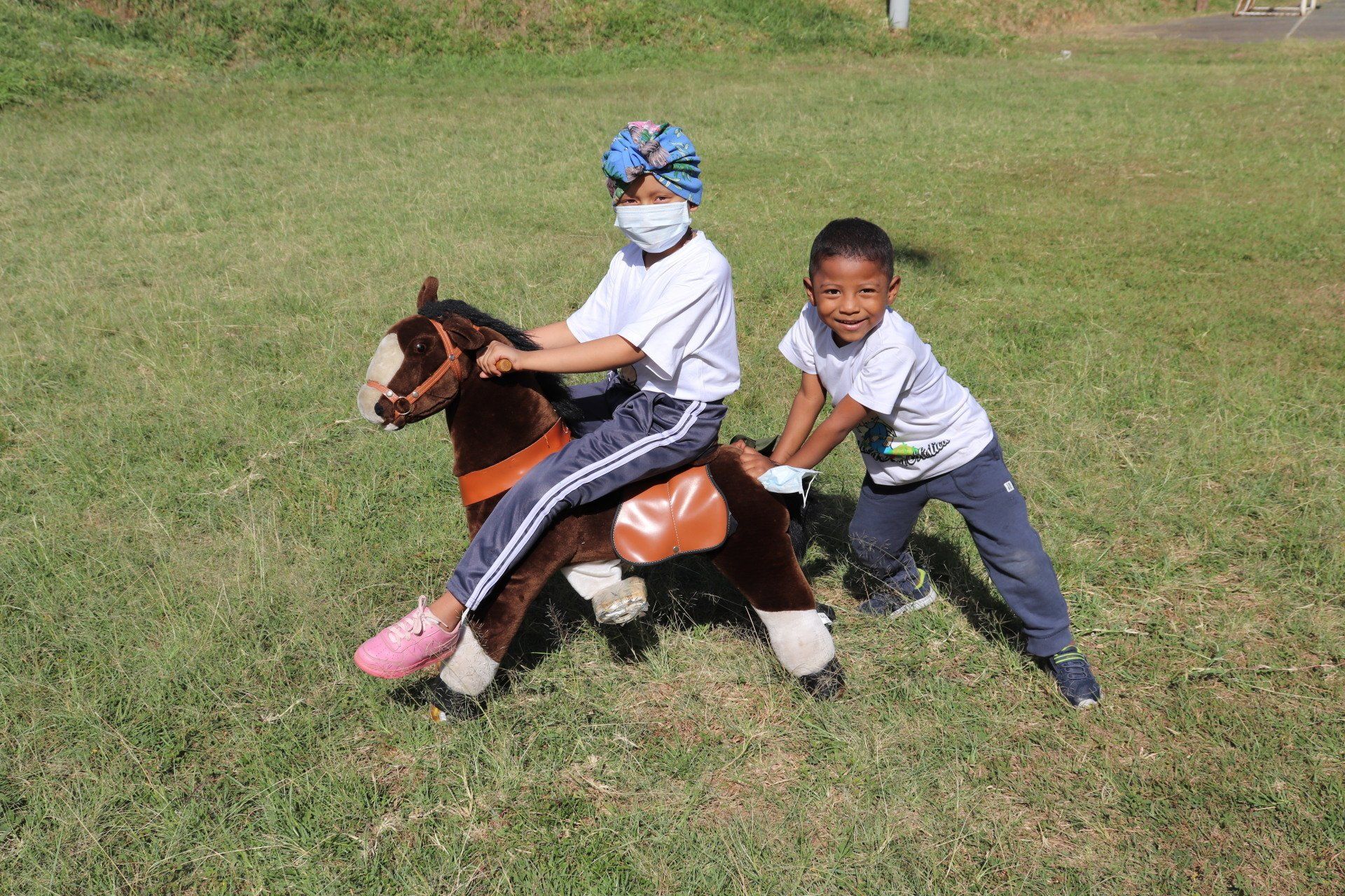 Dos niños jugando: uno monta un caballo de juguete, el otro lo tira sobre el césped.