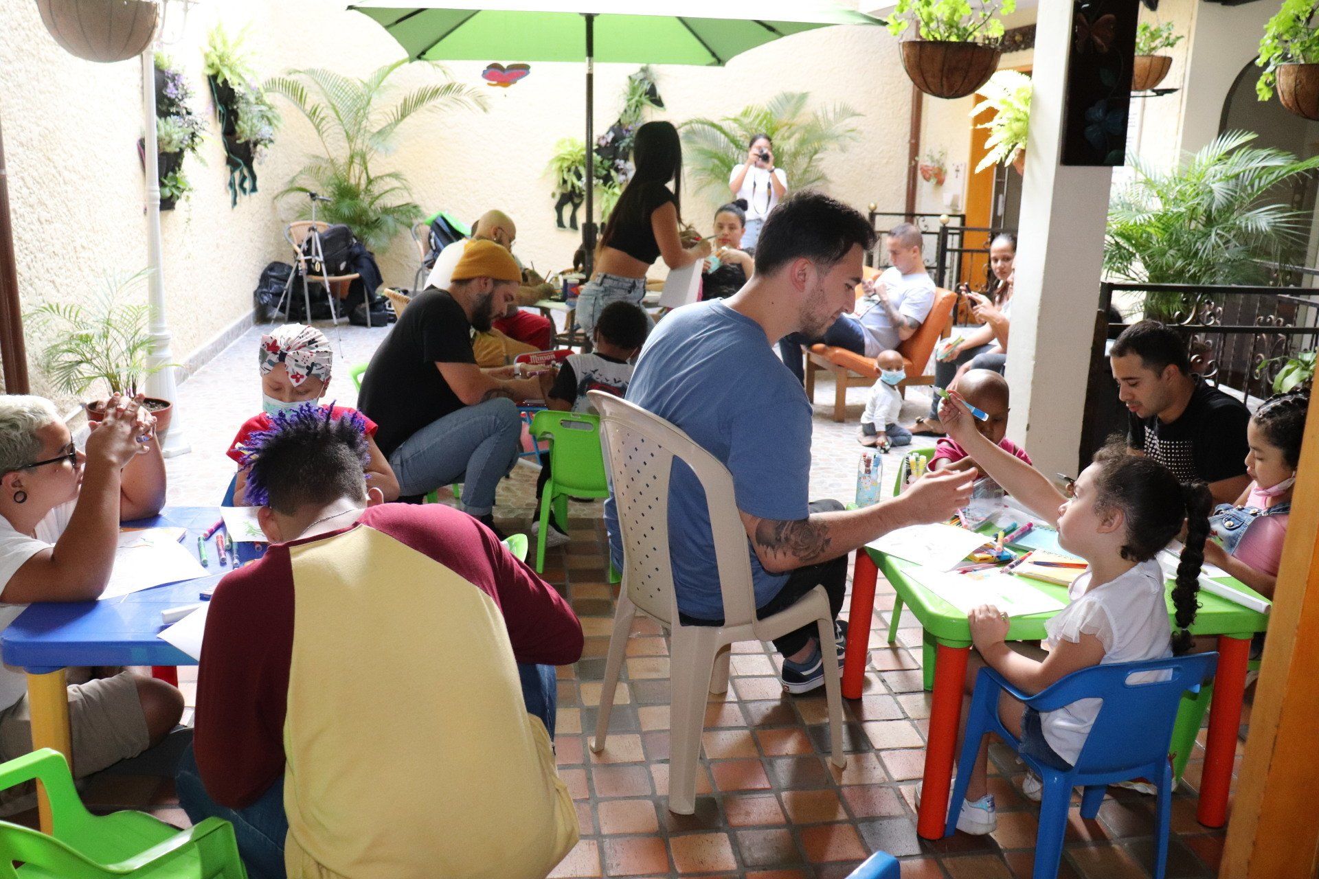 Personas sentadas en mesas pequeñas, coloreando con niños en un patio al aire libre con plantas.