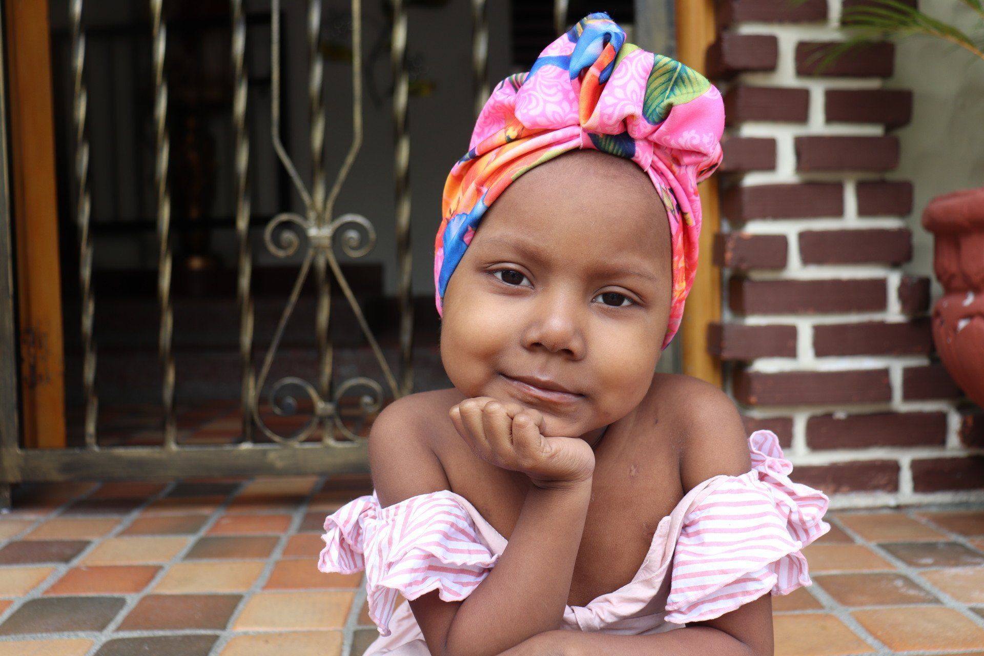 Niña con un colorido pañuelo en la cabeza y un vestido rosa, posando con la mano en la barbilla.