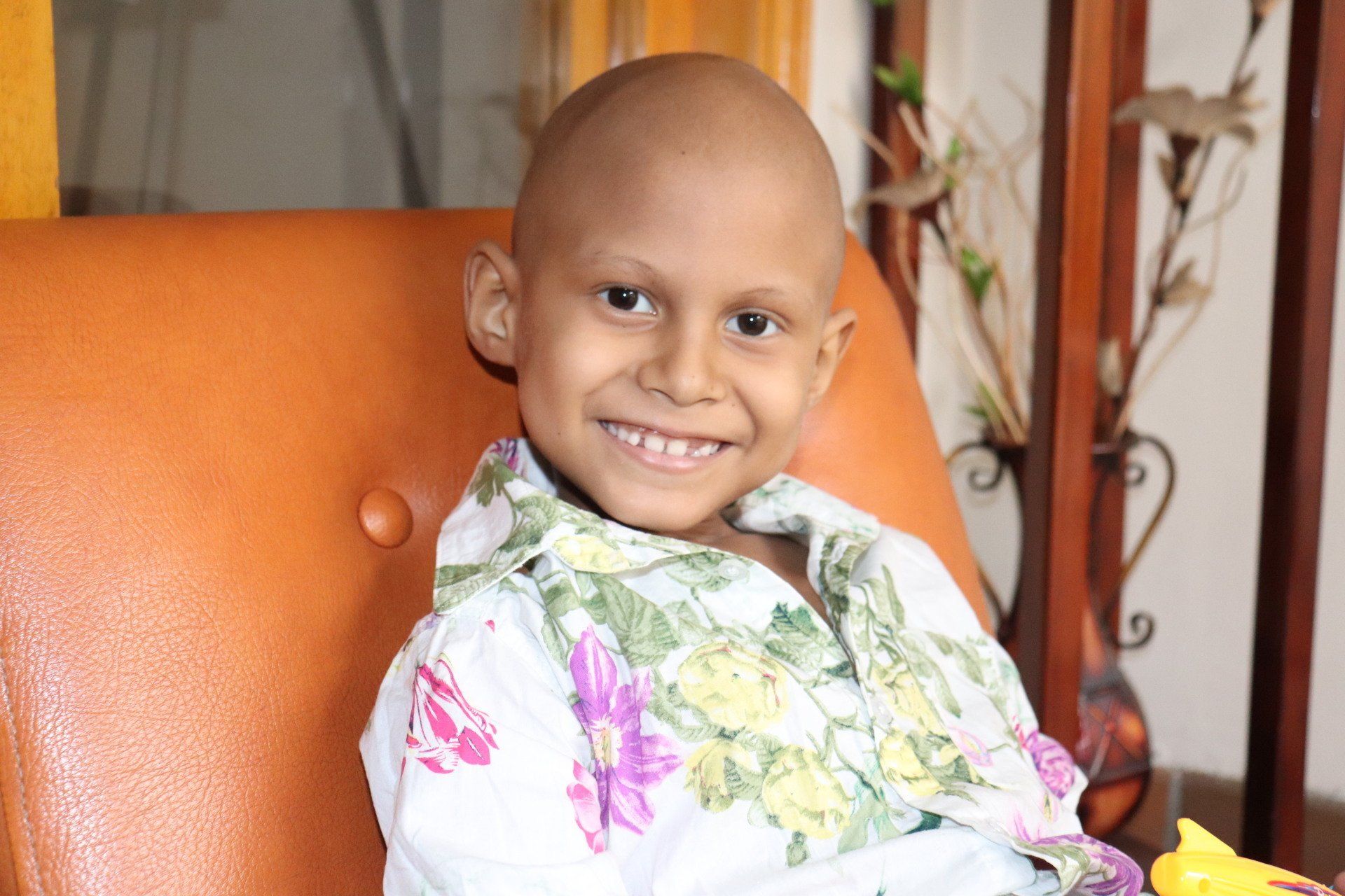 Un niño sonriente con la cabeza rapada se sienta en un sofá naranja y viste una camisa floreada.