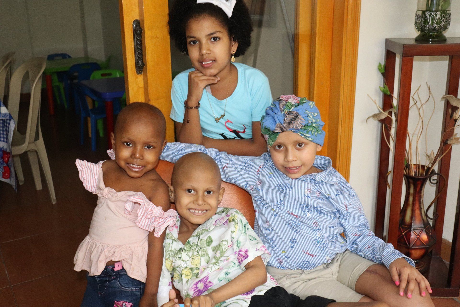 Cuatro niños, uno con diadema, tres con la cabeza rapada, sonriendo, sentados juntos, en un entorno interior.