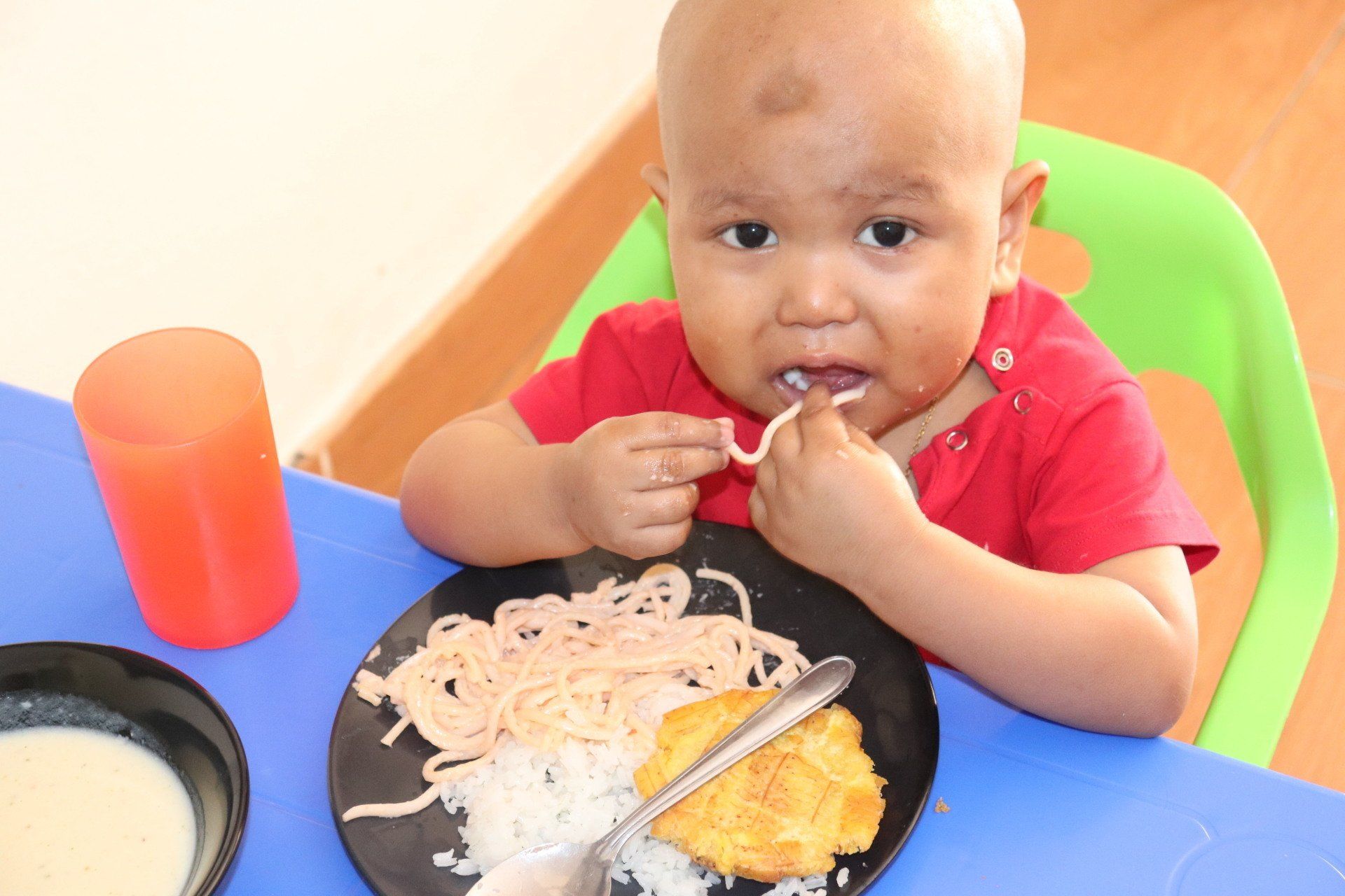 Un niño calvo con una camisa roja come pasta en una mesa con un plato negro y una taza naranja.
