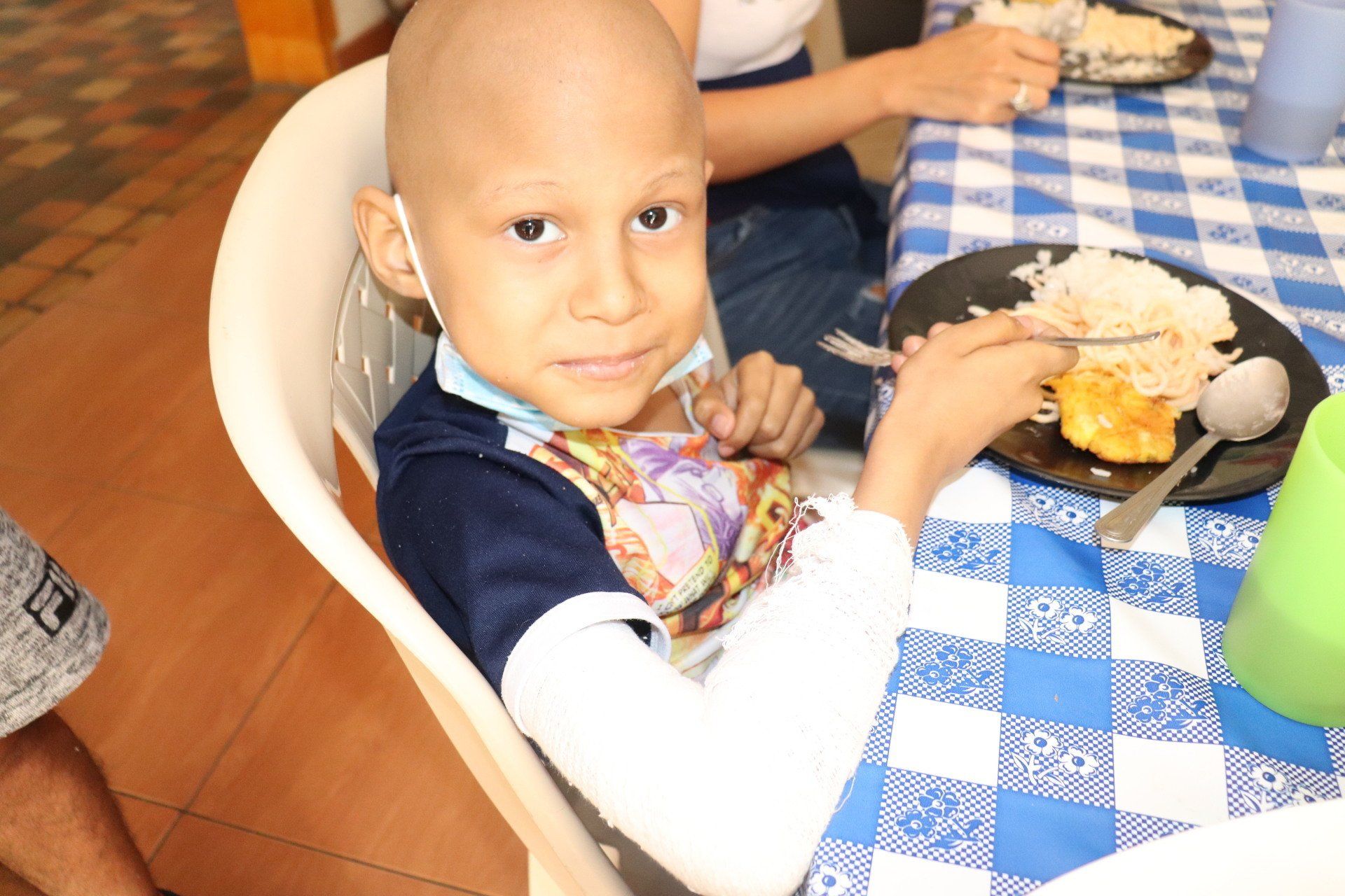Un niño con la cabeza rapada sonríe mientras come en una mesa a cuadros. 